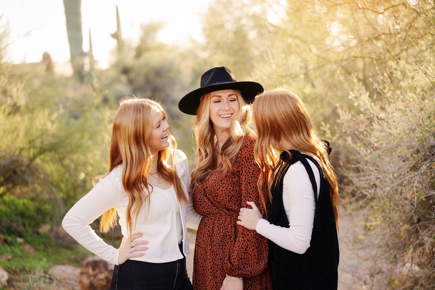 Three women laugh together on a desert trail with saguaro cacti and golden sunlight in the background.
