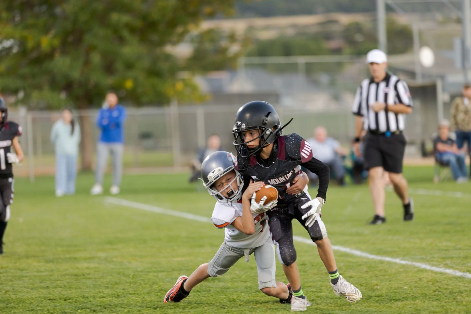 A lacrosse player in black uniform crouches low while defending against an opponent on a sunny grass field.