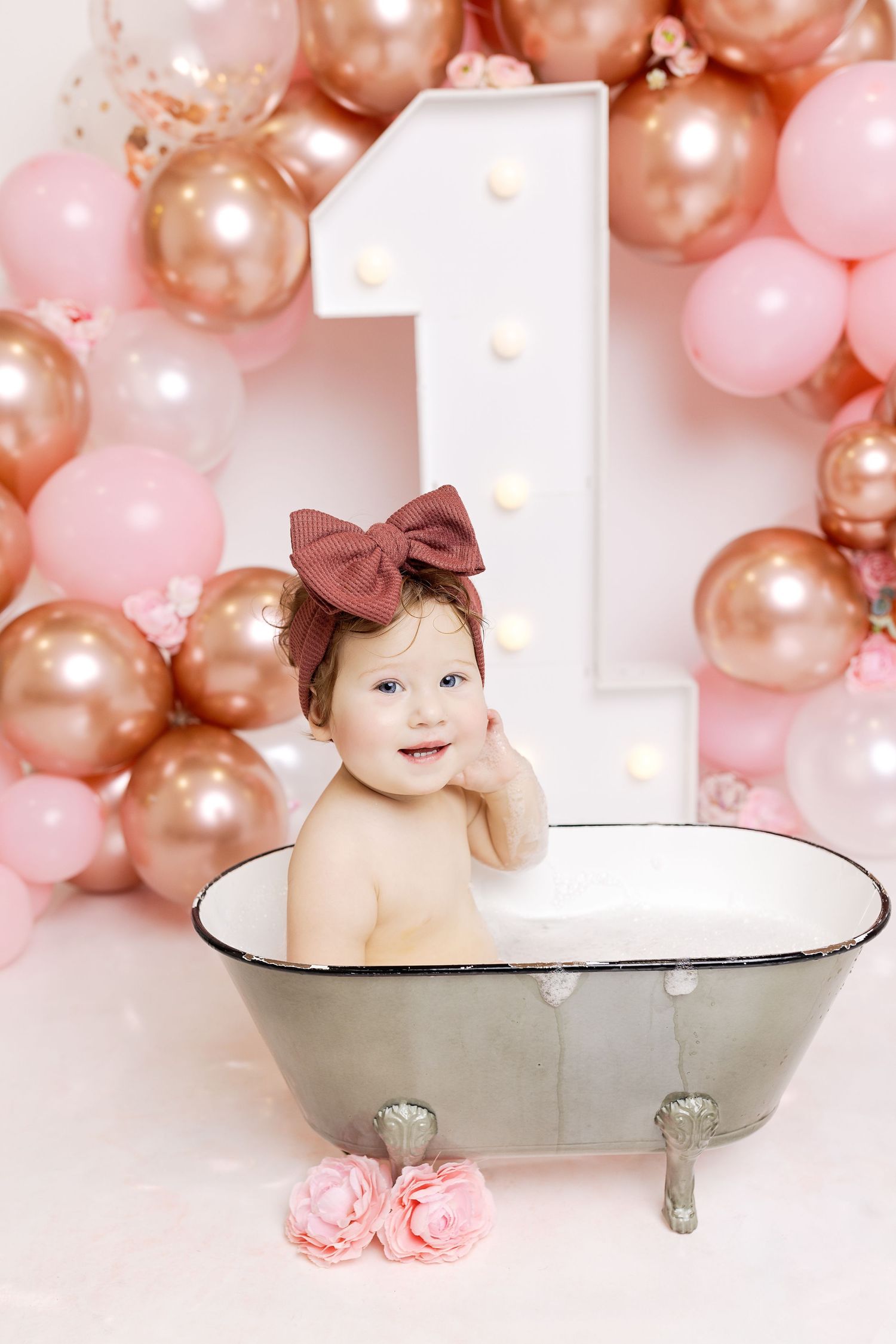 Pink and rose gold balloon garland decorates a vintage bathtub setup against a white backdrop.