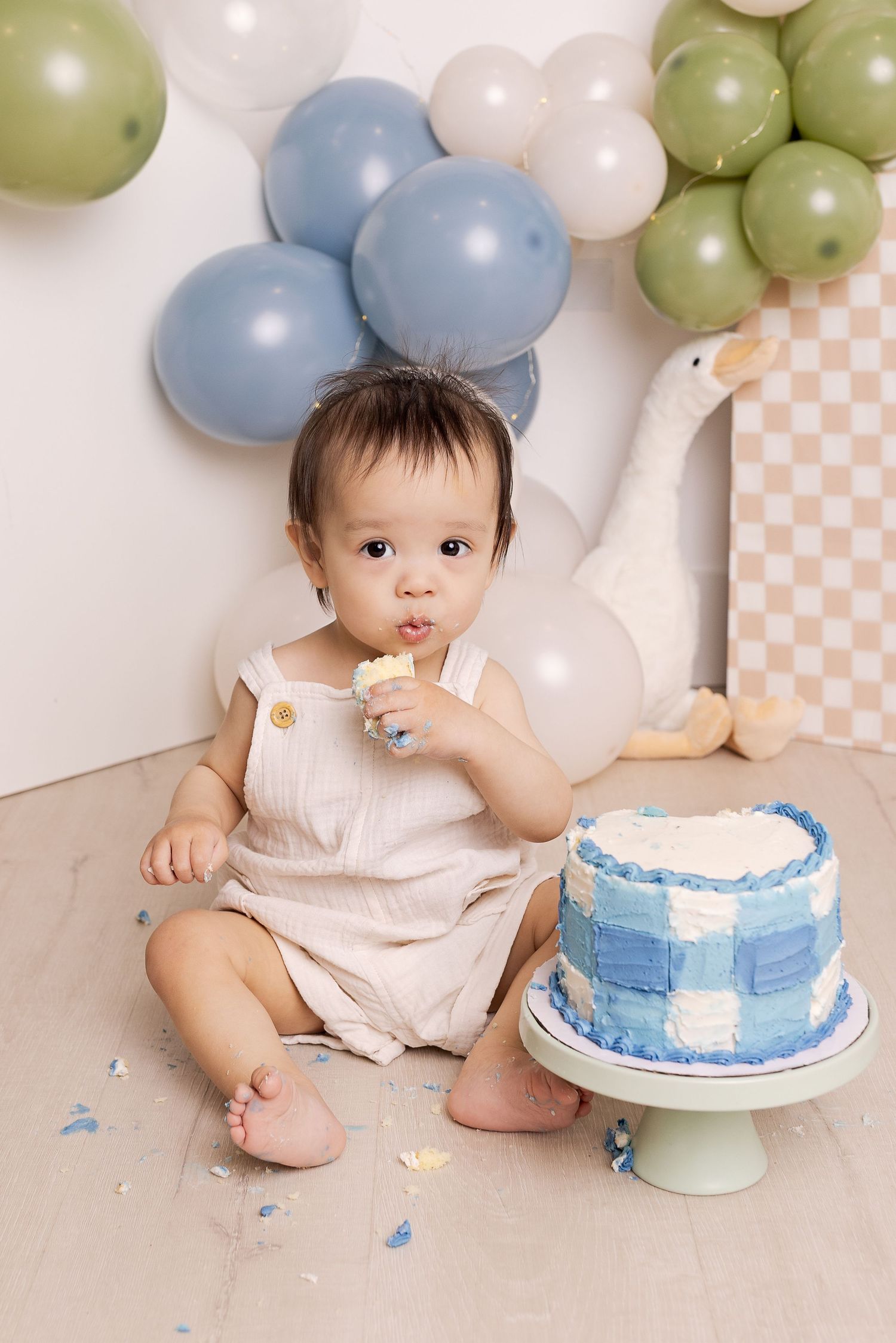 Baby in linen romper enjoys first birthday cake smash with blue gingham cake and balloon garland backdrop.