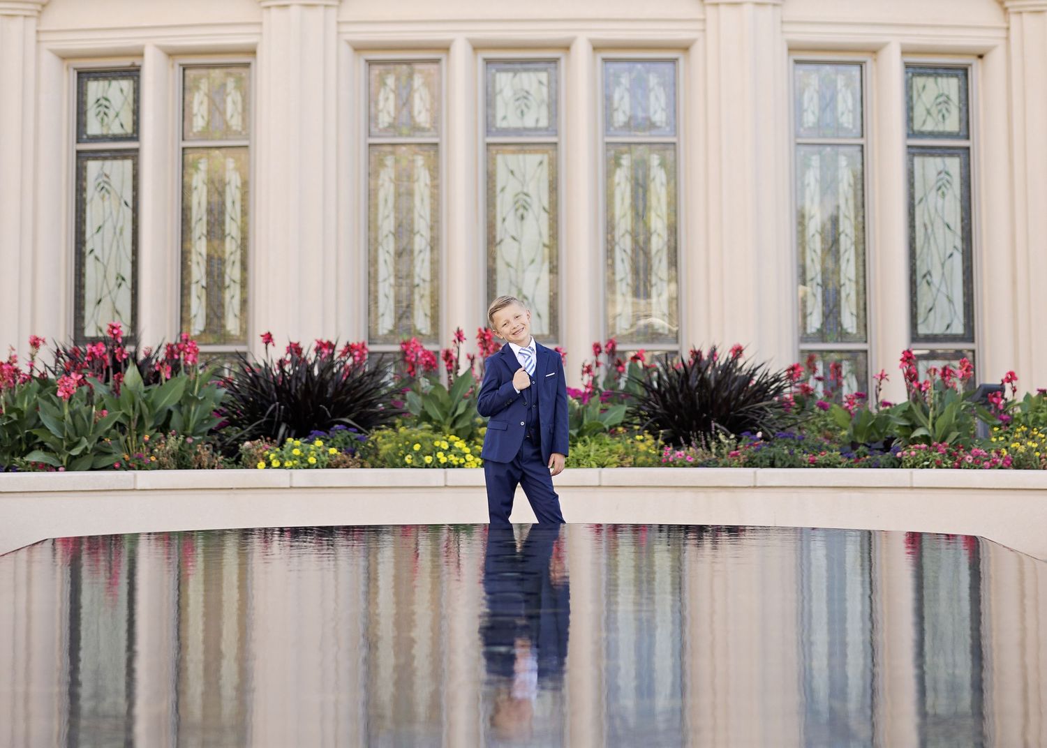 A figure in a navy suit stands reflected in a pool near ornate windows and pink flowers.