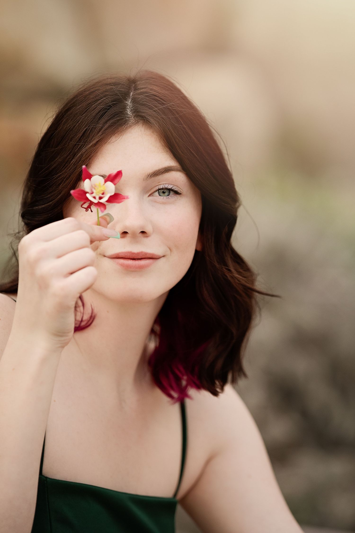 Artistic portrait with small red flower held up to face against blurred background.
