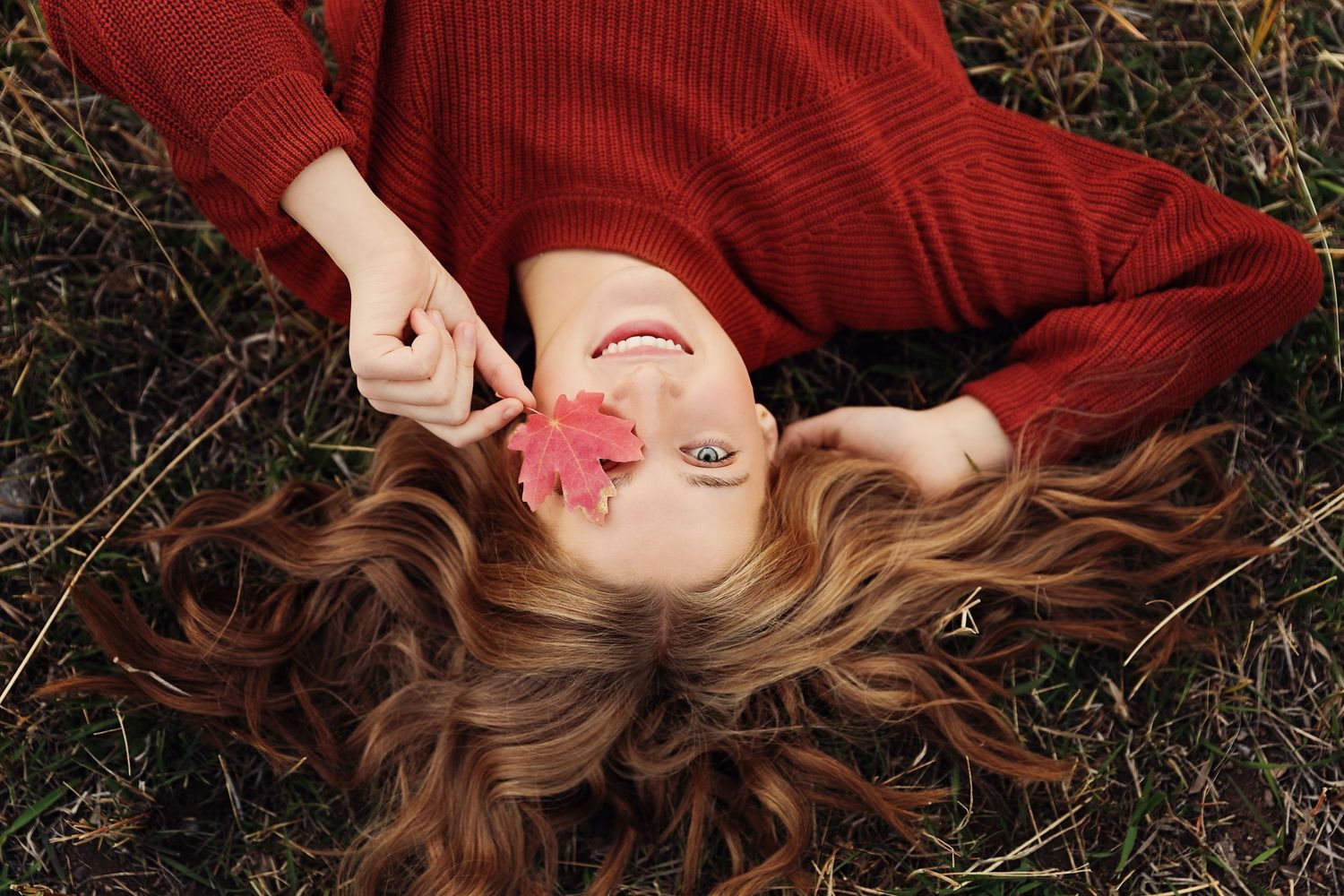 Creative portrait lying in grass wearing red sweater with long wavy hair spread out.
