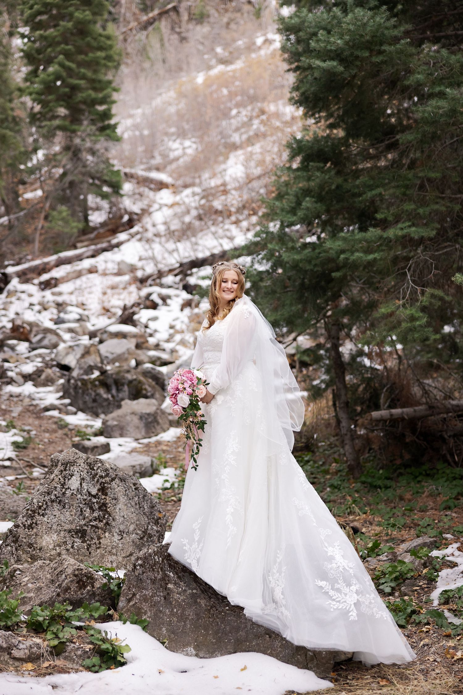 Bride with flowing veil and pink bouquet poses on snowy mountain trail near evergreen trees.