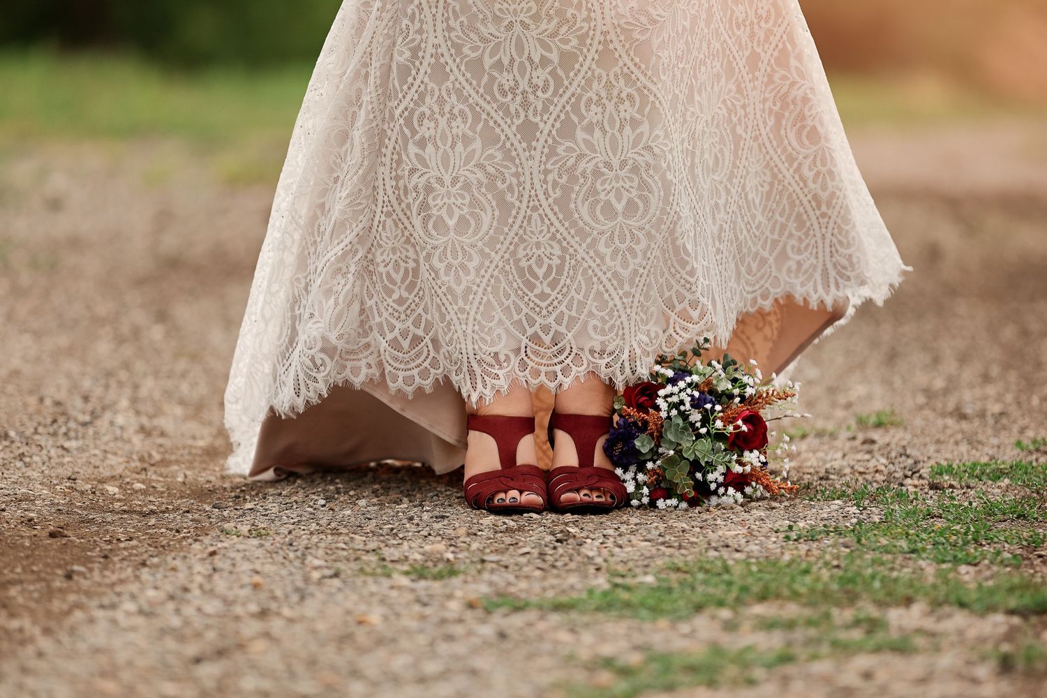 Creative wedding detail shot showing red shoes and bouquet beneath lace dress hem.