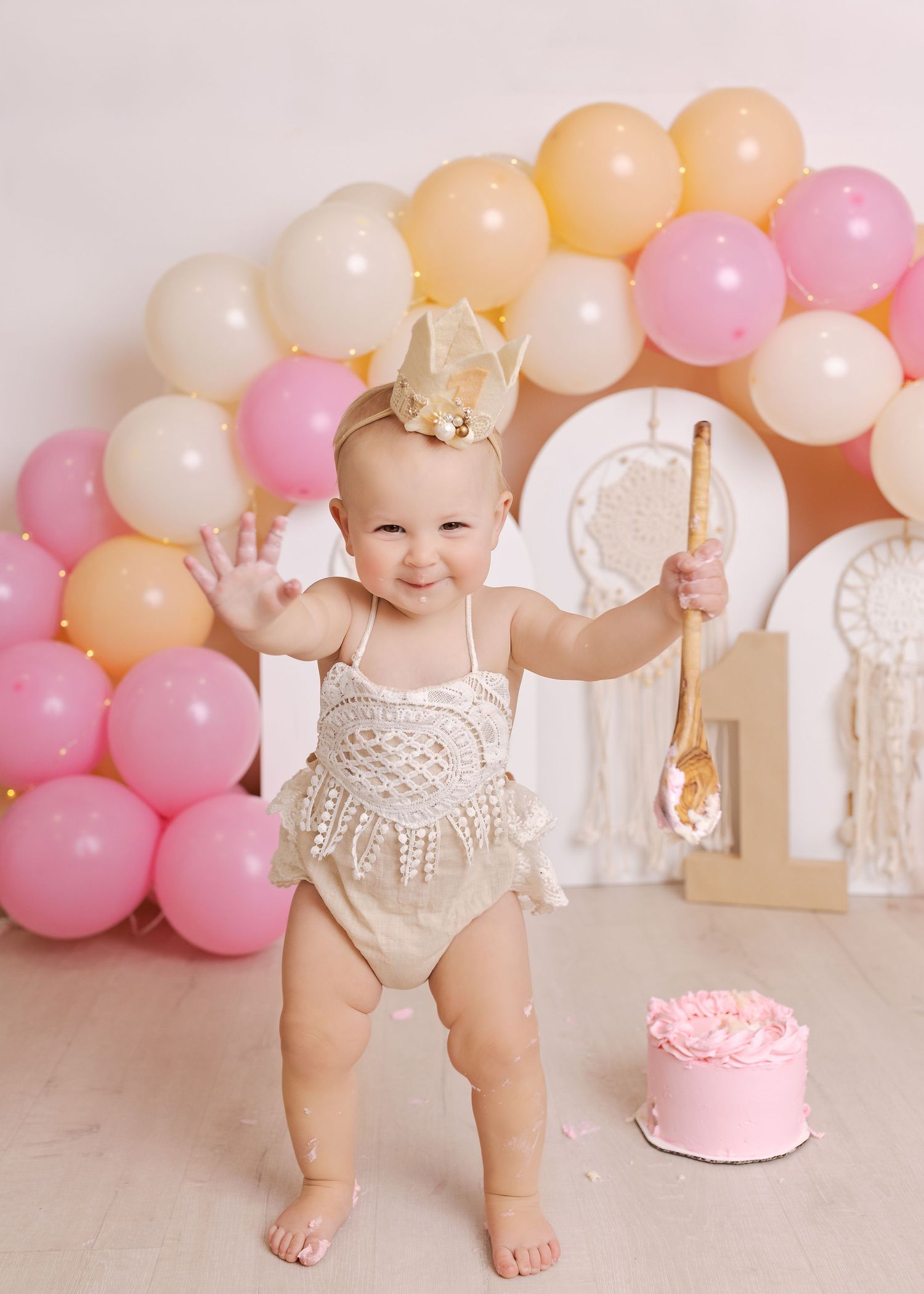 Pink and peach balloon garland decorates first birthday cake smash photo session with white backdrop and sweet details.