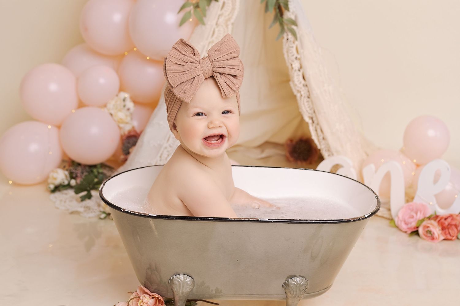 Baby in vintage washtub surrounded by pink balloons and floral decorations during cake smash photo session.