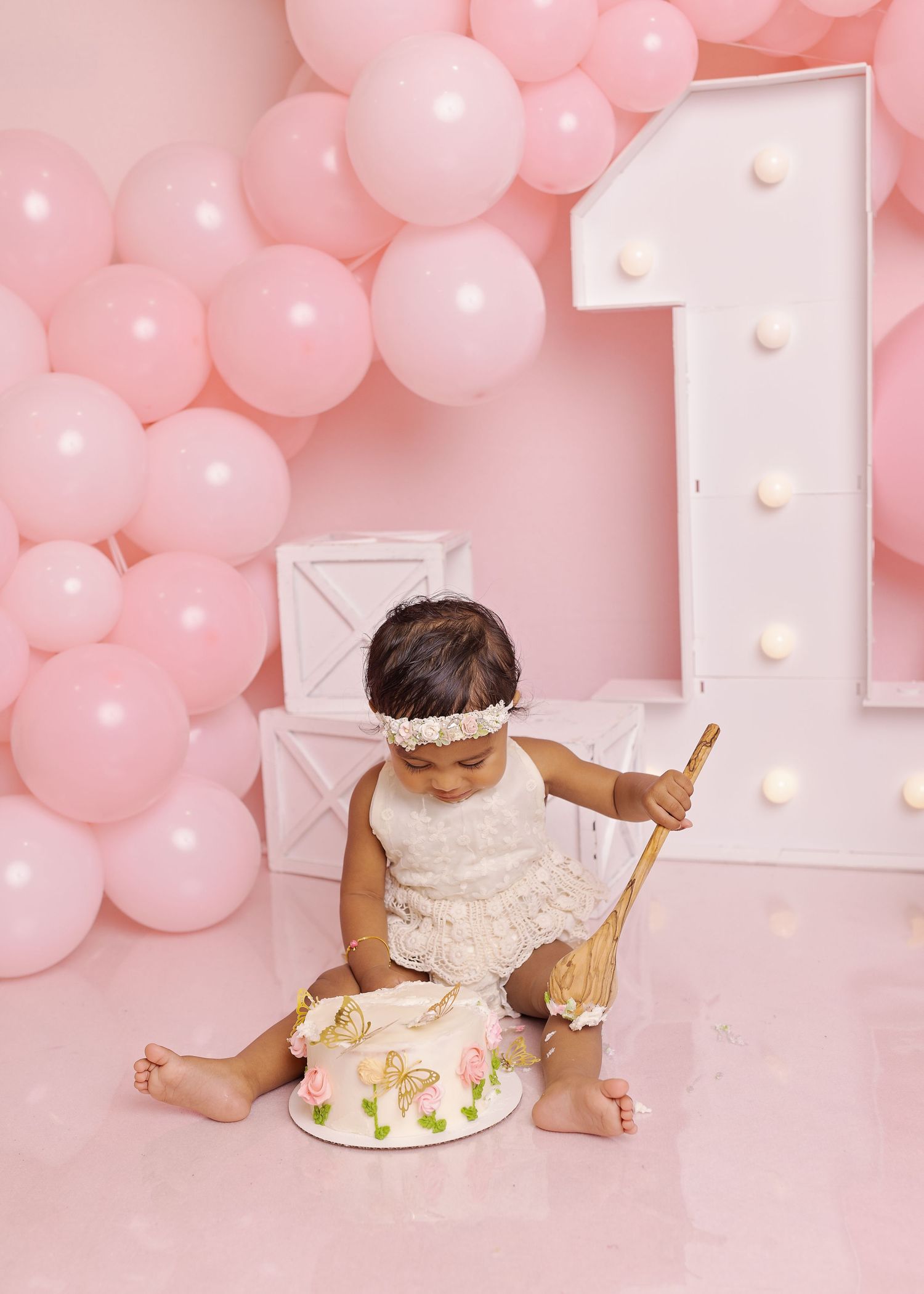 A baby sits on a pink backdrop with balloons and a light-up number 1, celebrating their first birthday with a cake.