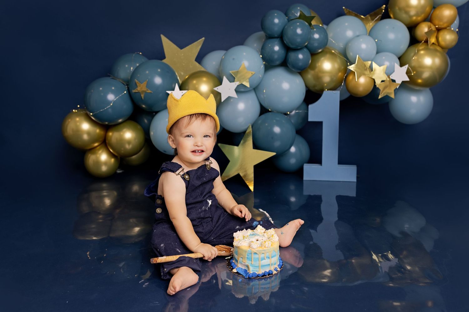 First birthday cake smash photo with blue and gold balloon arch and star decorations against dark backdrop.
