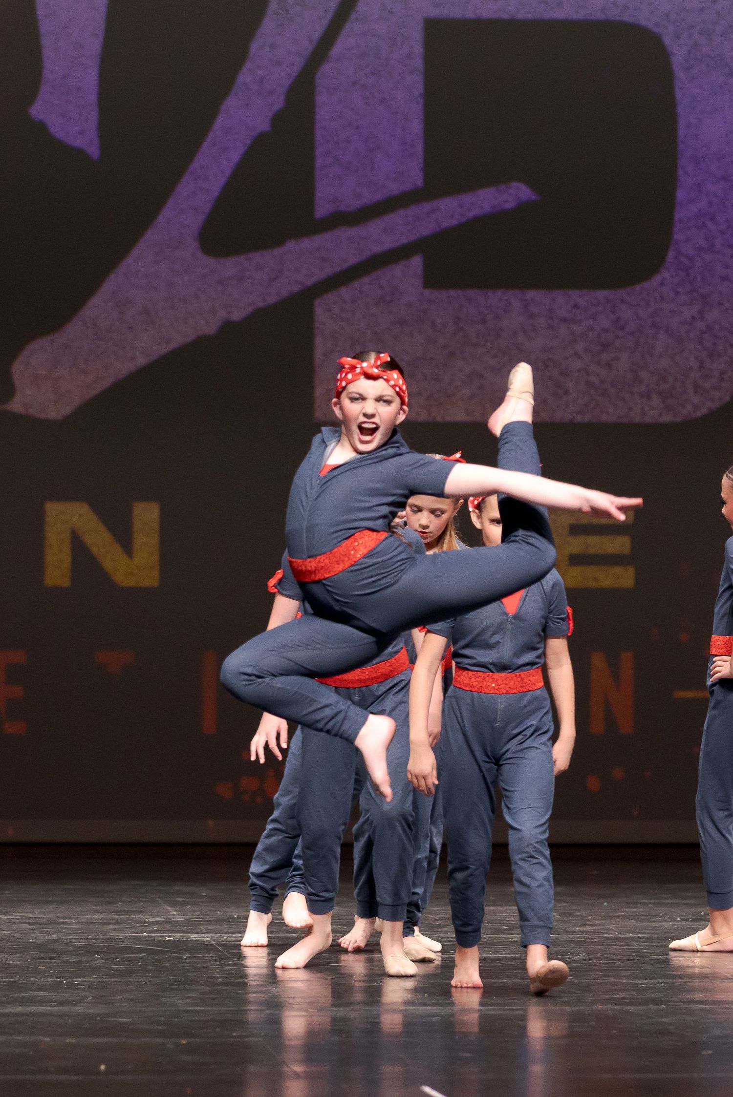 Group of dancers in navy blue outfits and red belts perform dynamic contemporary dance routine on dark stage.