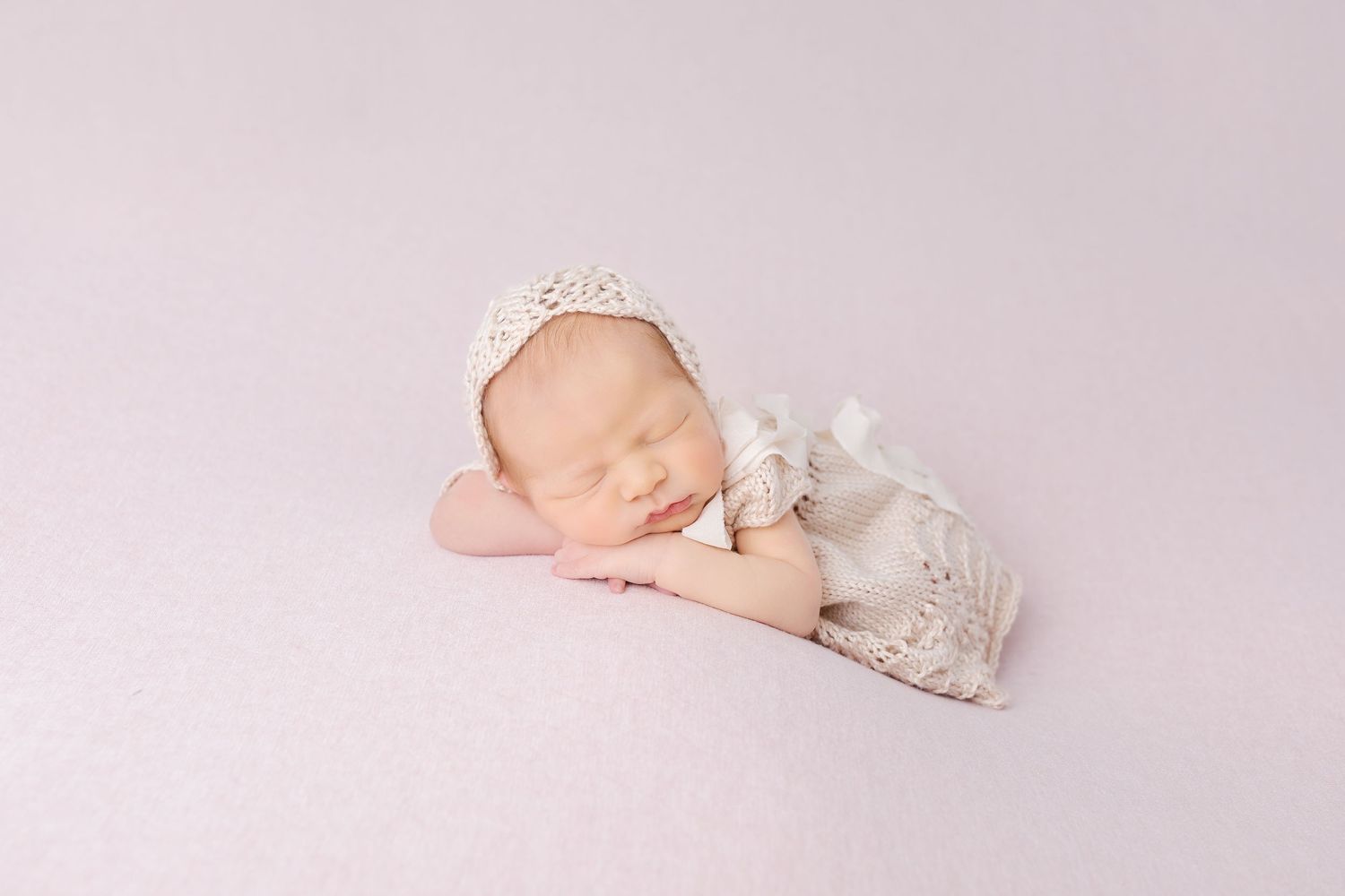 Peaceful newborn baby sleeping in cream lace outfit on soft pink background.