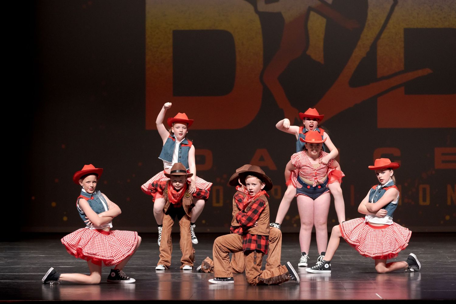 Performers in western-themed costumes dance on stage during a theatrical production with rustic backdrop.