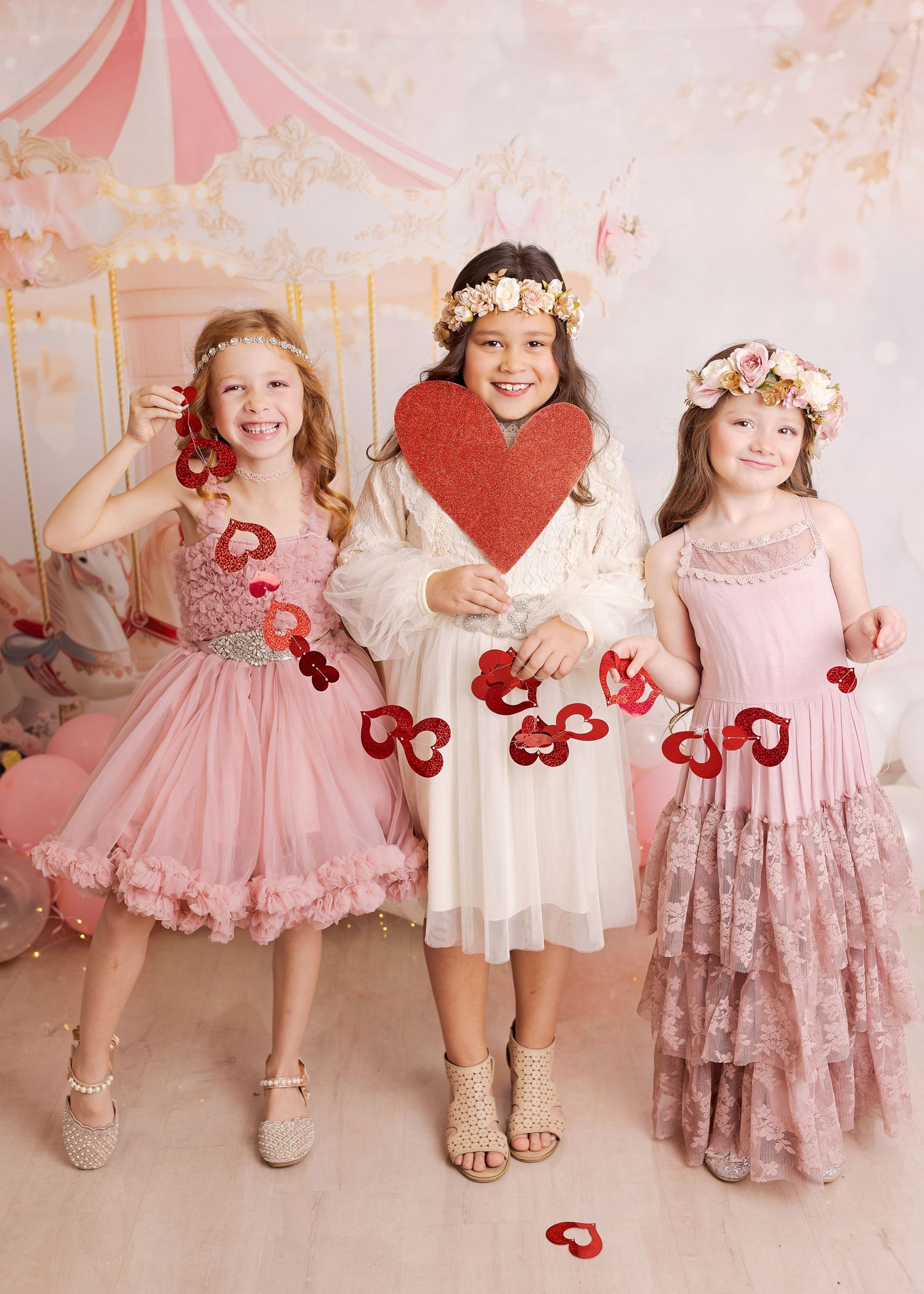 A group of children wearing pink and white dresses with tulle skirts stand together against a pink backdrop with heart decorations.