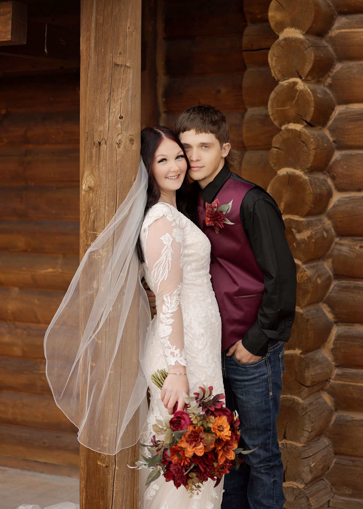 Romantic wedding portrait against rustic log cabin backdrop with burgundy and orange floral accents.