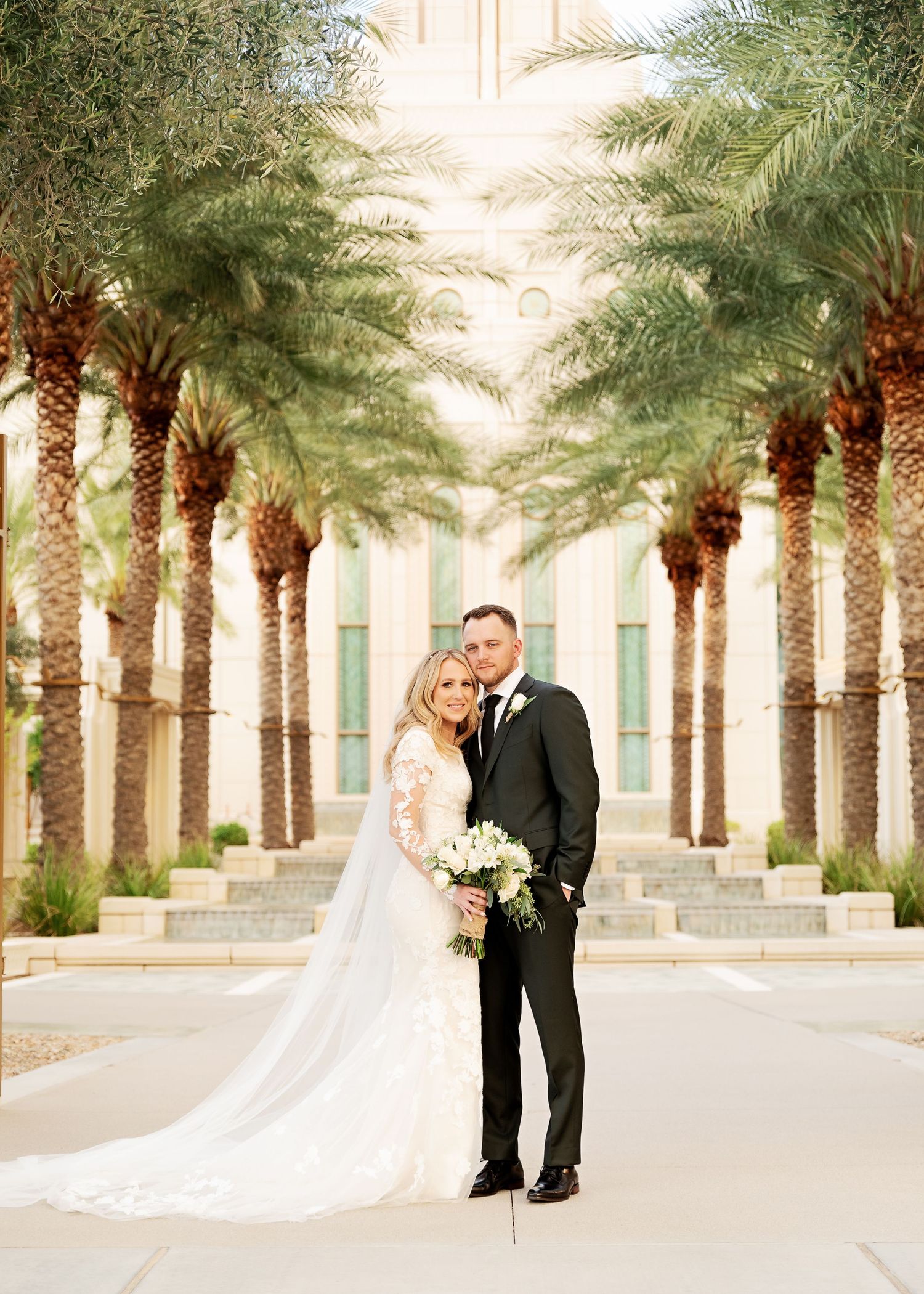 Wedding couple poses between rows of palm trees on pathway.