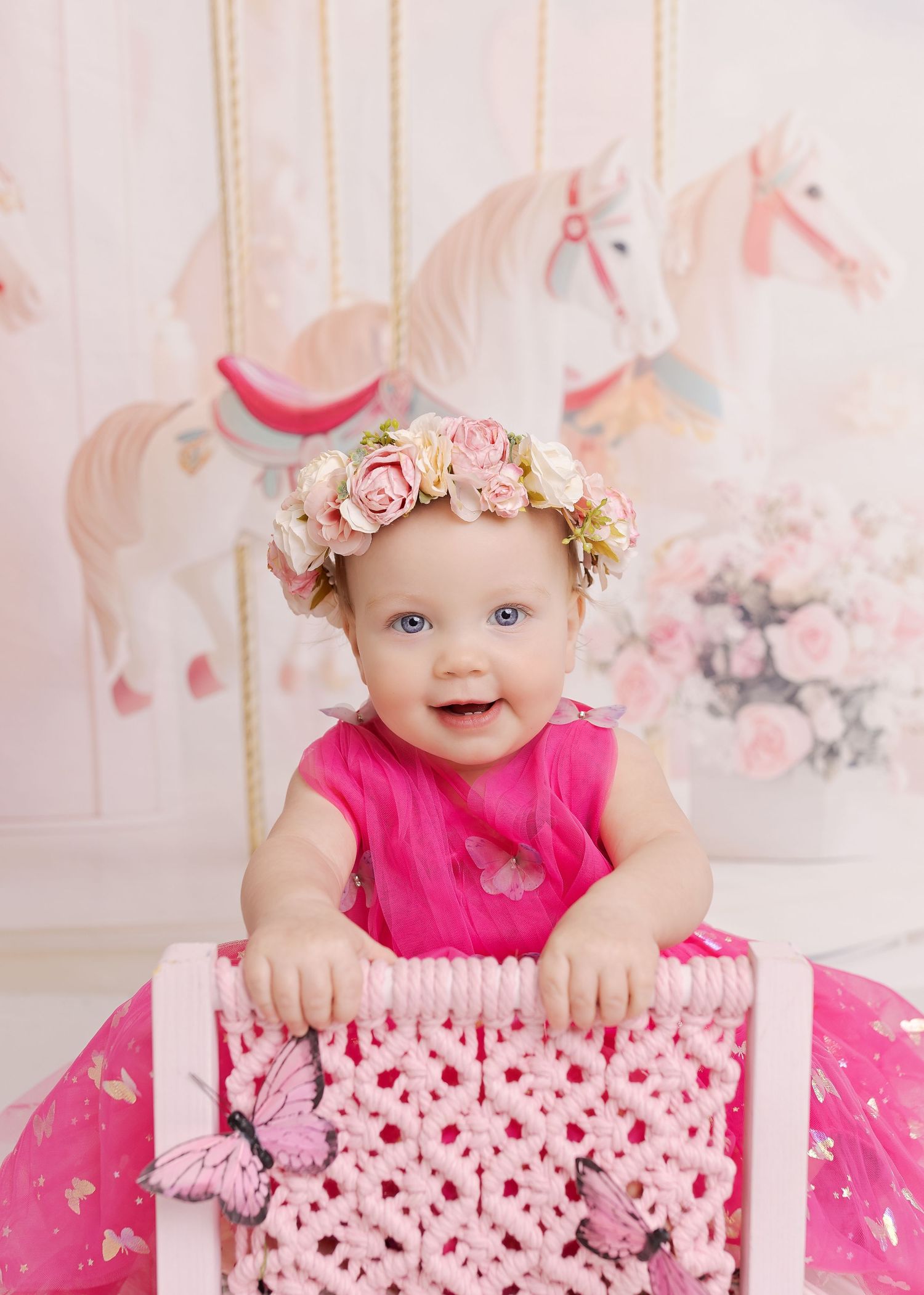 Baby in pink dress wearing floral crown poses against carousel horse themed backdrop with pink decor.