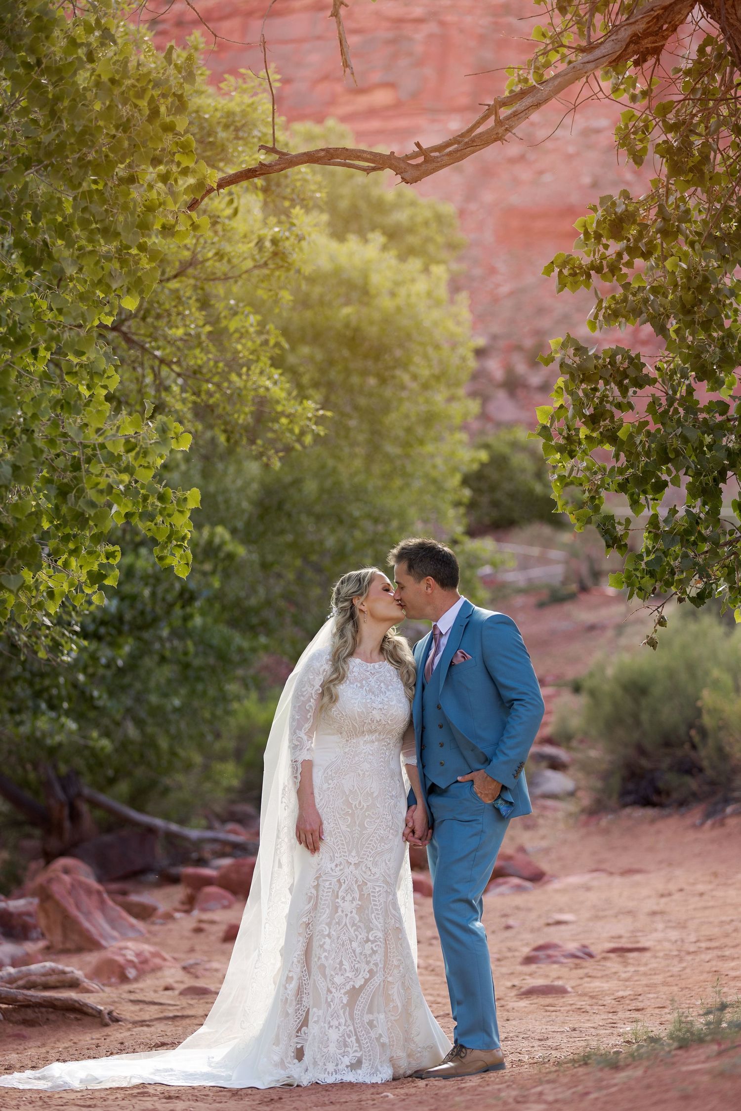 A wedding couple shares romantic moments in a majestic red rock canyon with desert greenery at sunset.