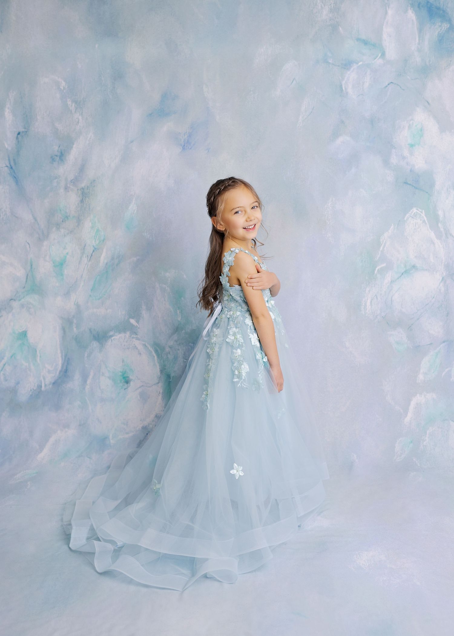 A dreamy portrait of a child in a flowing light blue tulle gown with floral details against a soft blue background.