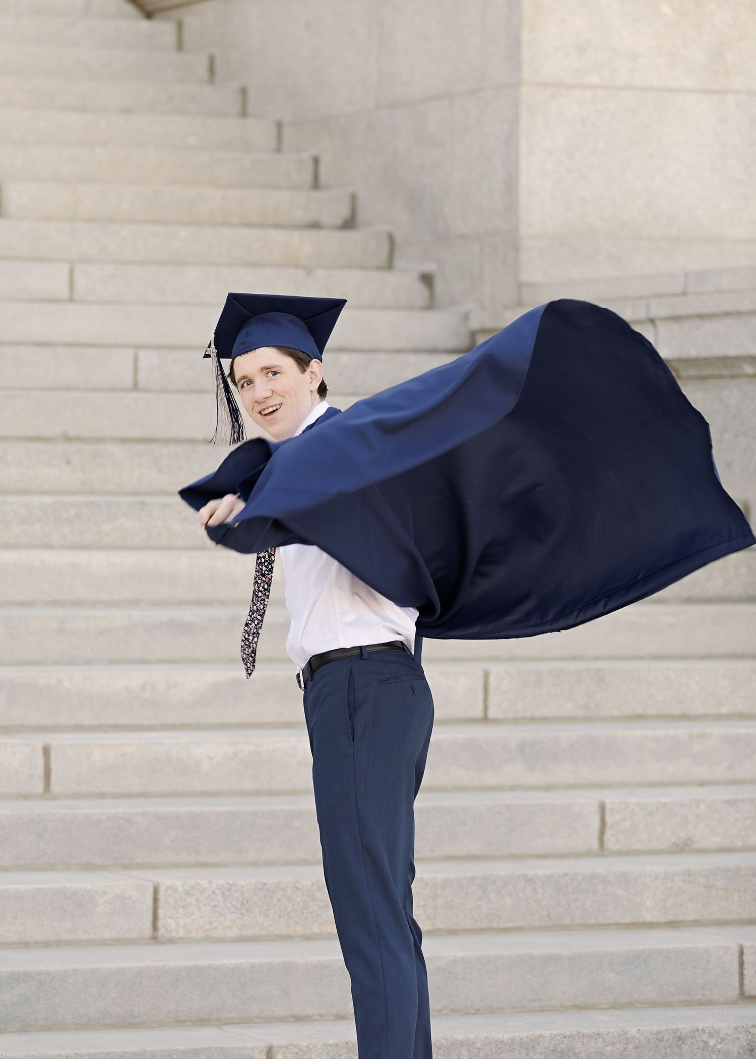 Graduate in navy cap and gown posing dramatically on white stone steps.