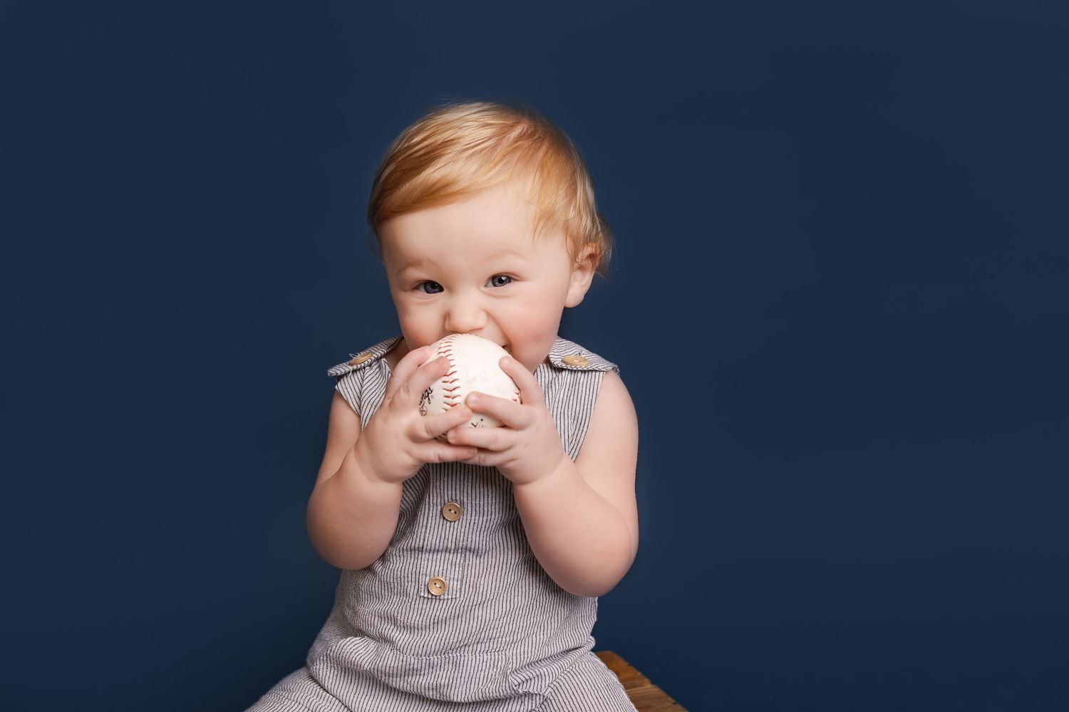 Young child in gray outfit bringing baseball up to mouth against navy background.