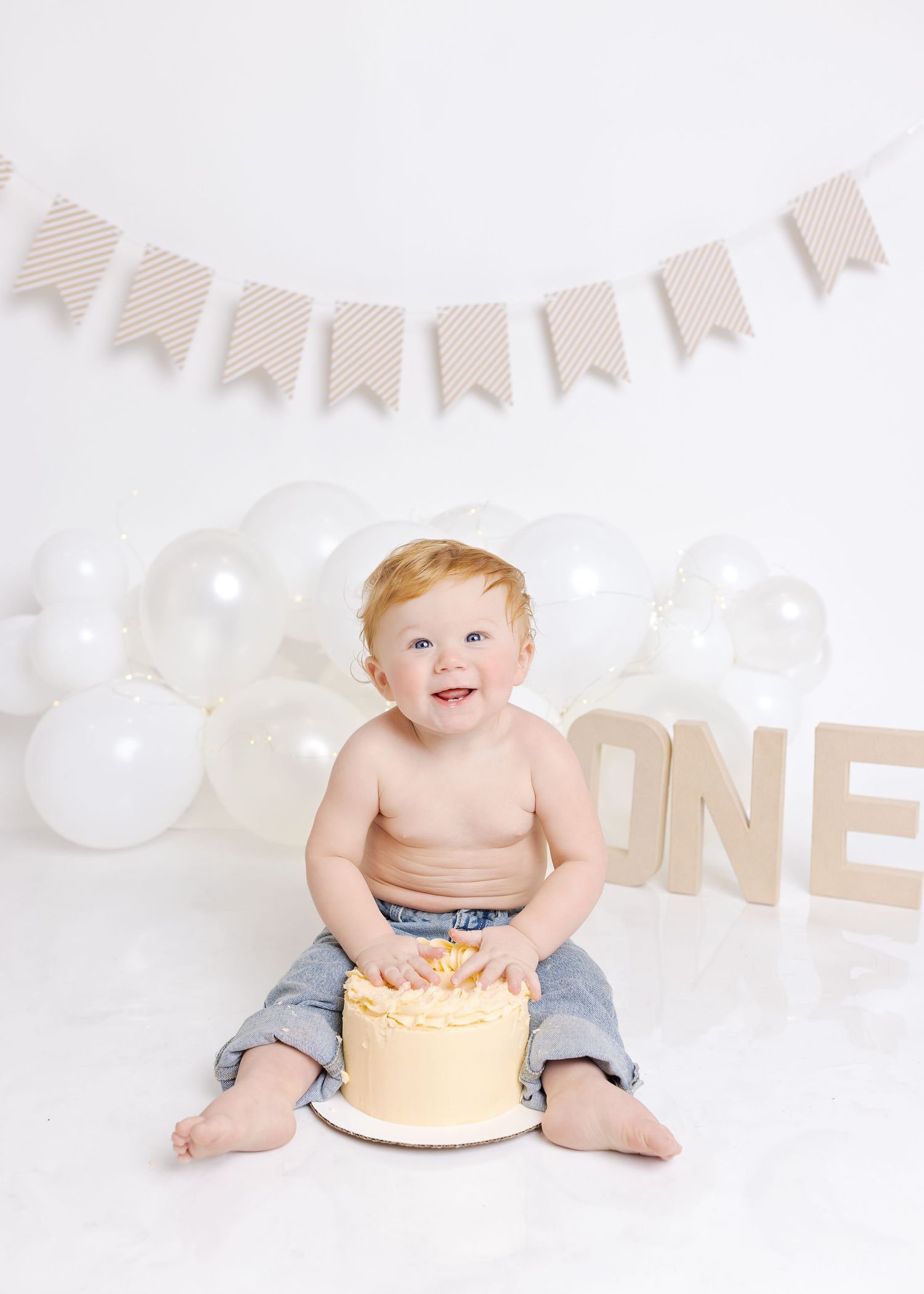 Baby wearing denim shorts sits with birthday cake for first birthday celebration with neutral bunting decoration above.