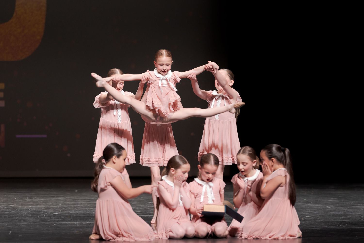 Dancers in matching pink dresses perform an intricate choreographed formation on a dark stage during a recital.