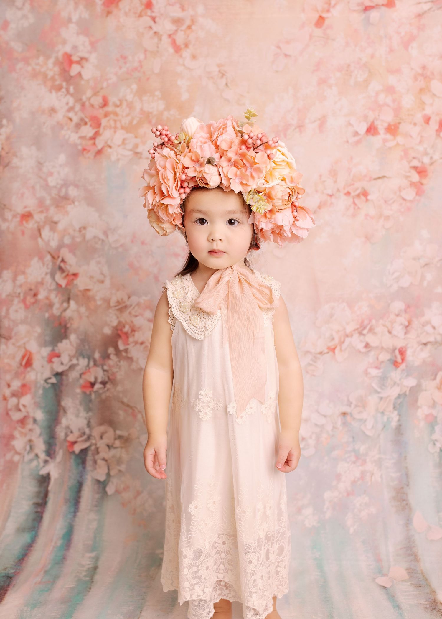 Child wearing lace dress and pink floral crown against soft pink backdrop.