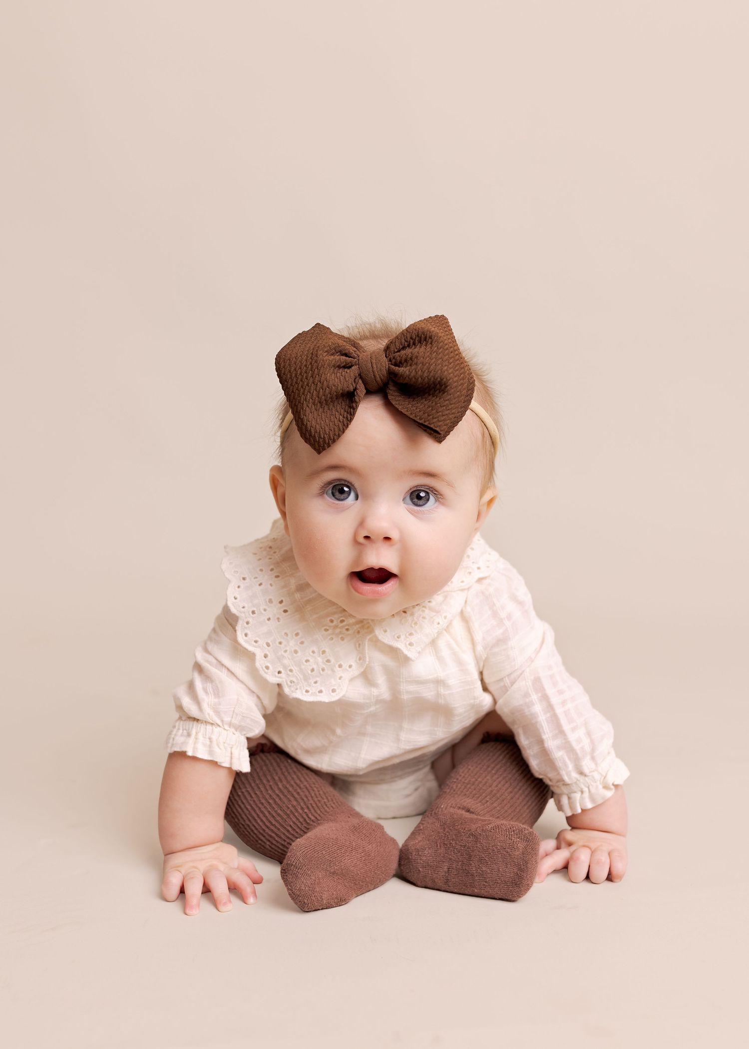 A baby wearing a brown bow and pink dress lies on their tummy on a cream-colored background.