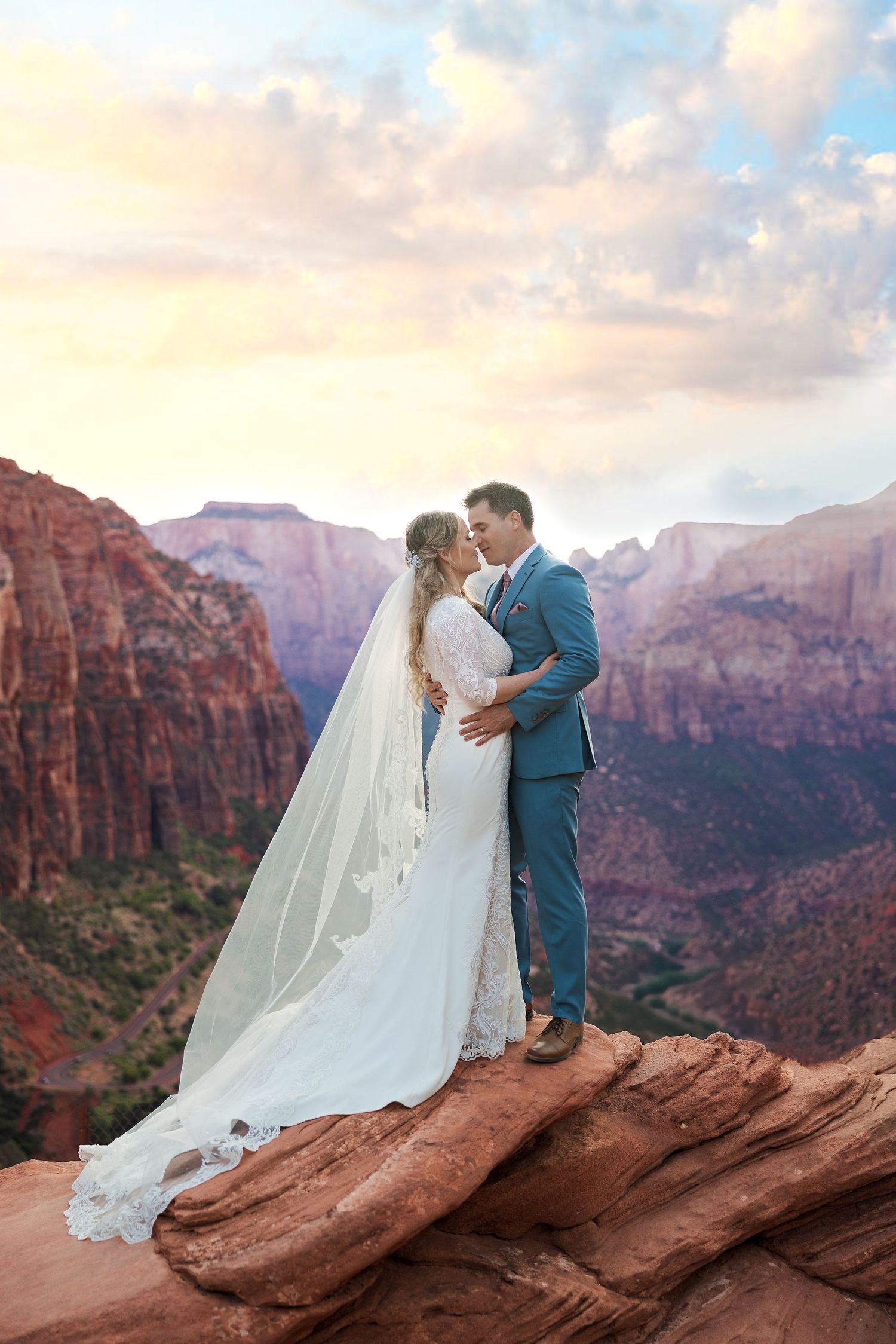 A couple stands atop red rocks in Zion National Park during sunset with dramatic mountain landscape in the background.