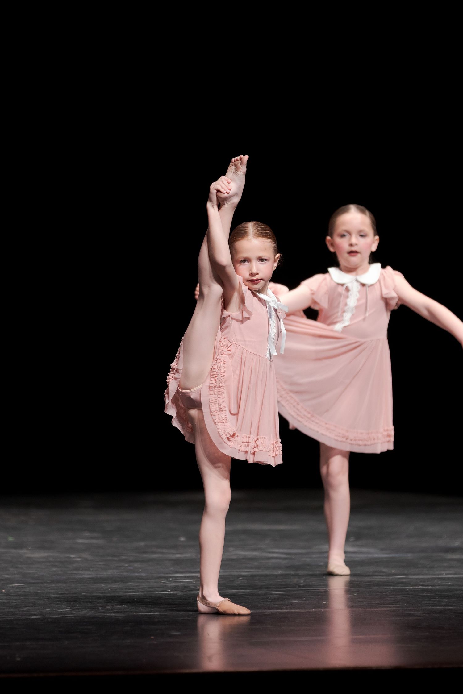 Two young dancers in pink dresses perform graceful ballet moves on a dark stage.