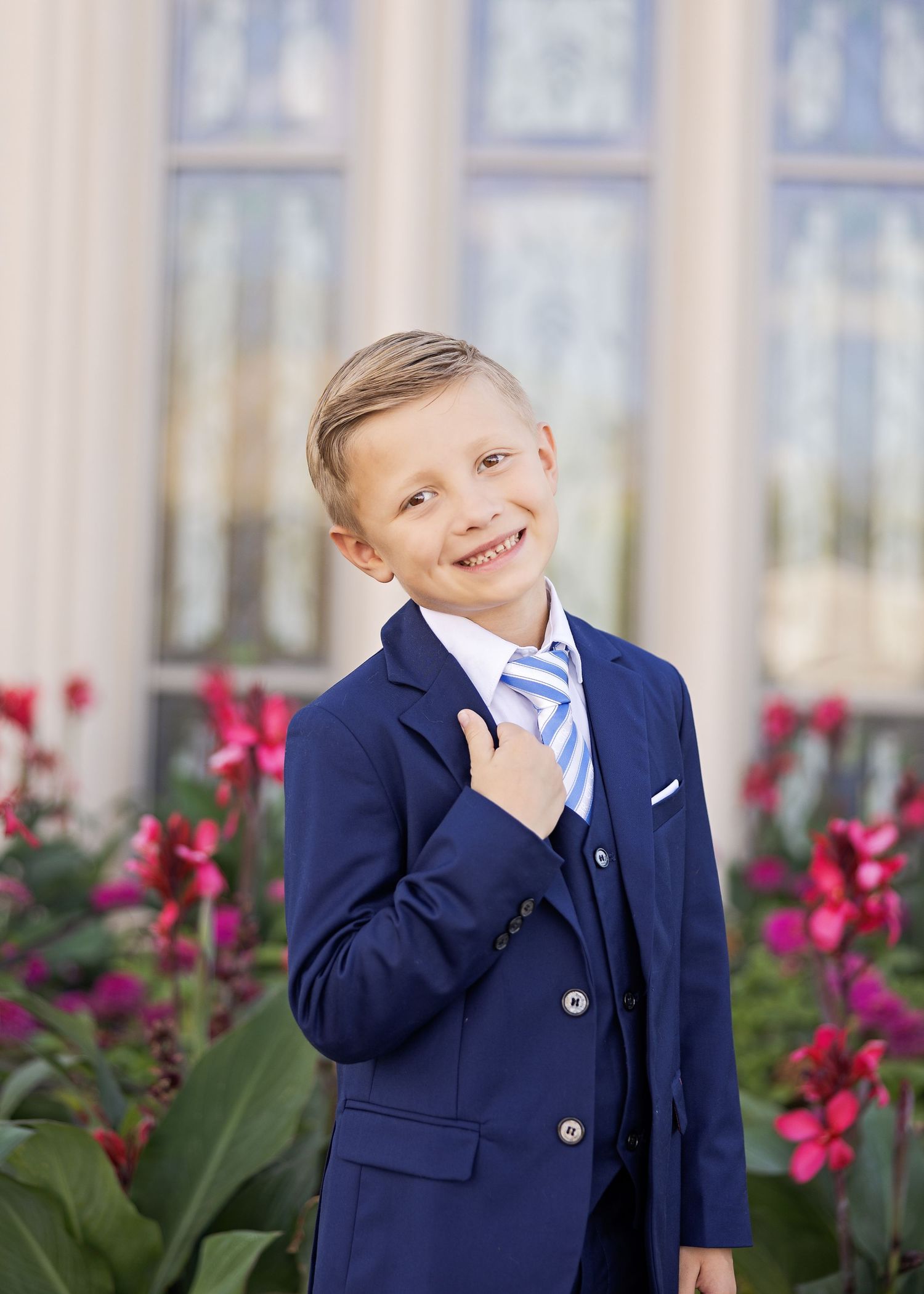 A young person in a navy blue suit and striped tie poses outdoors near pink flowers and ornate windows.