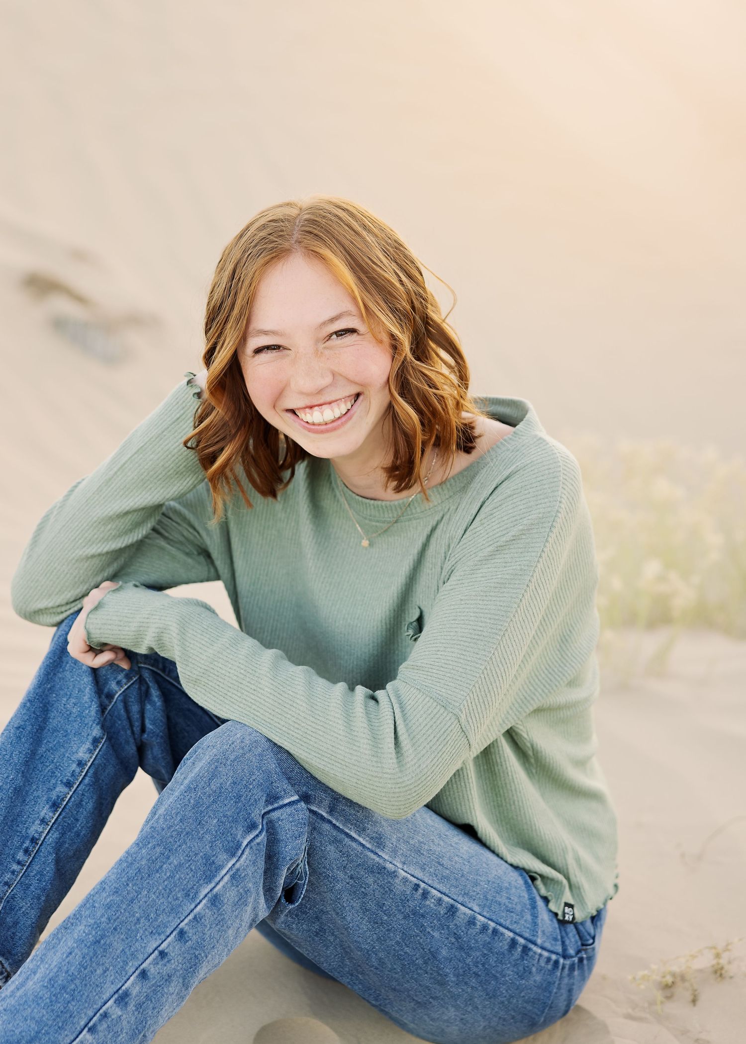 A casual portrait of someone in a sage green sweater and jeans sitting outdoors.