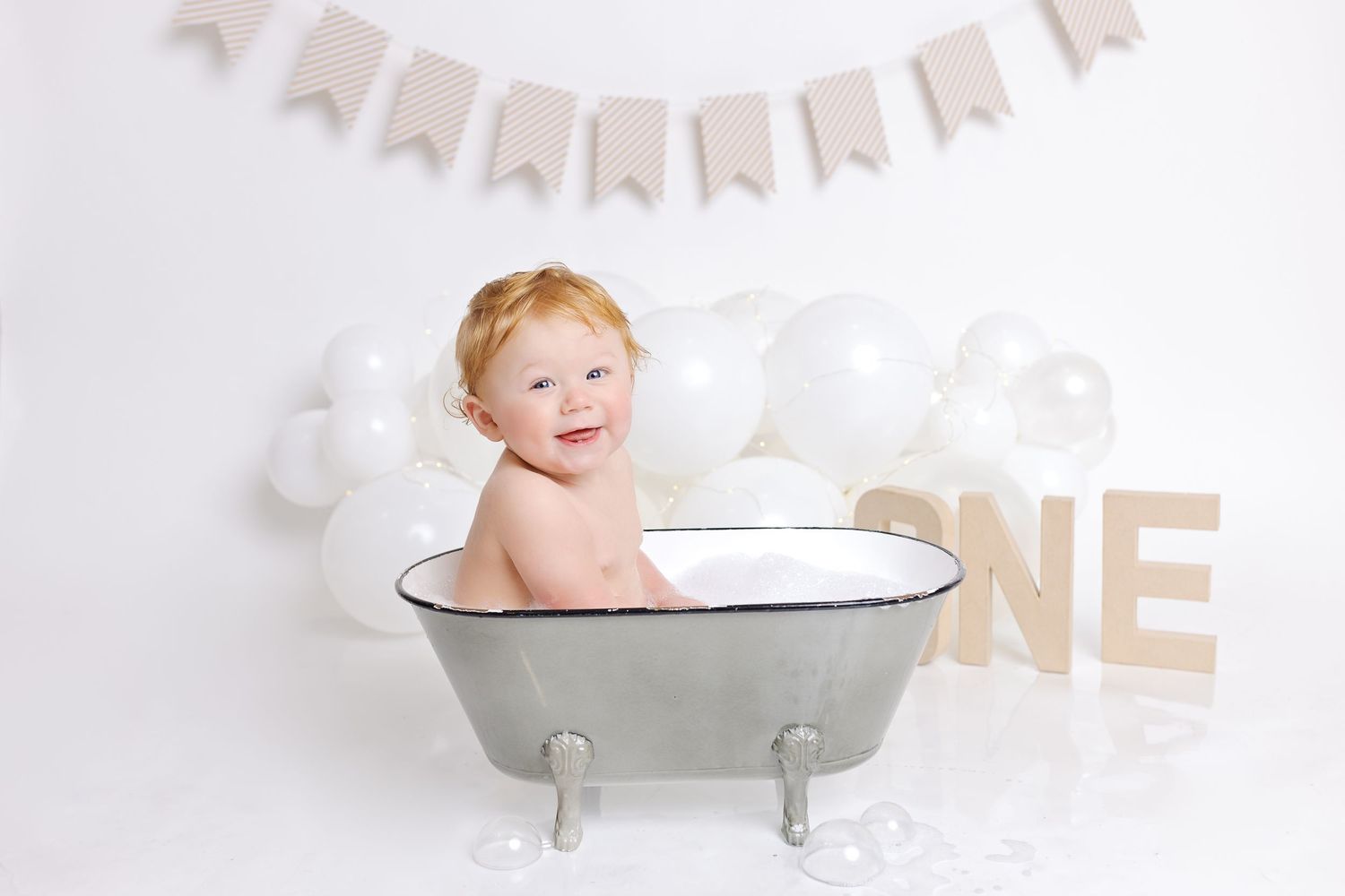 A baby sits in a white miniature clawfoot bathtub under a banner for a first birthday photoshoot.