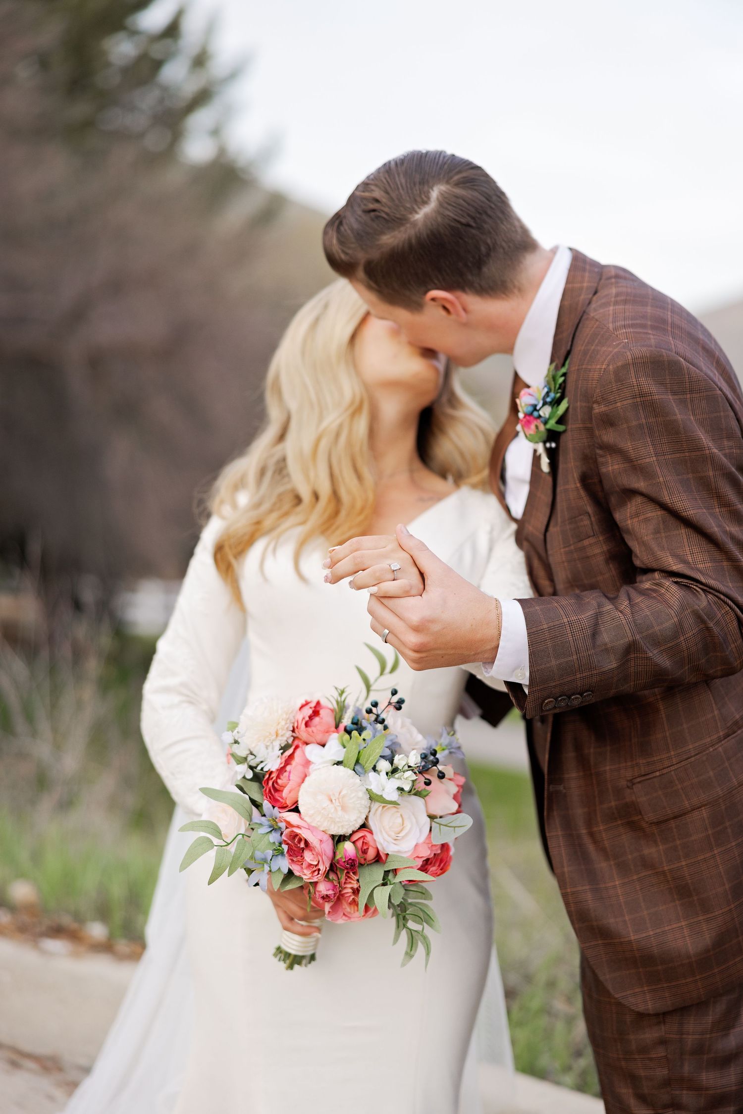 A romantic wedding couple shares a tender moment while holding a pink and coral floral bouquet in an outdoor setting.