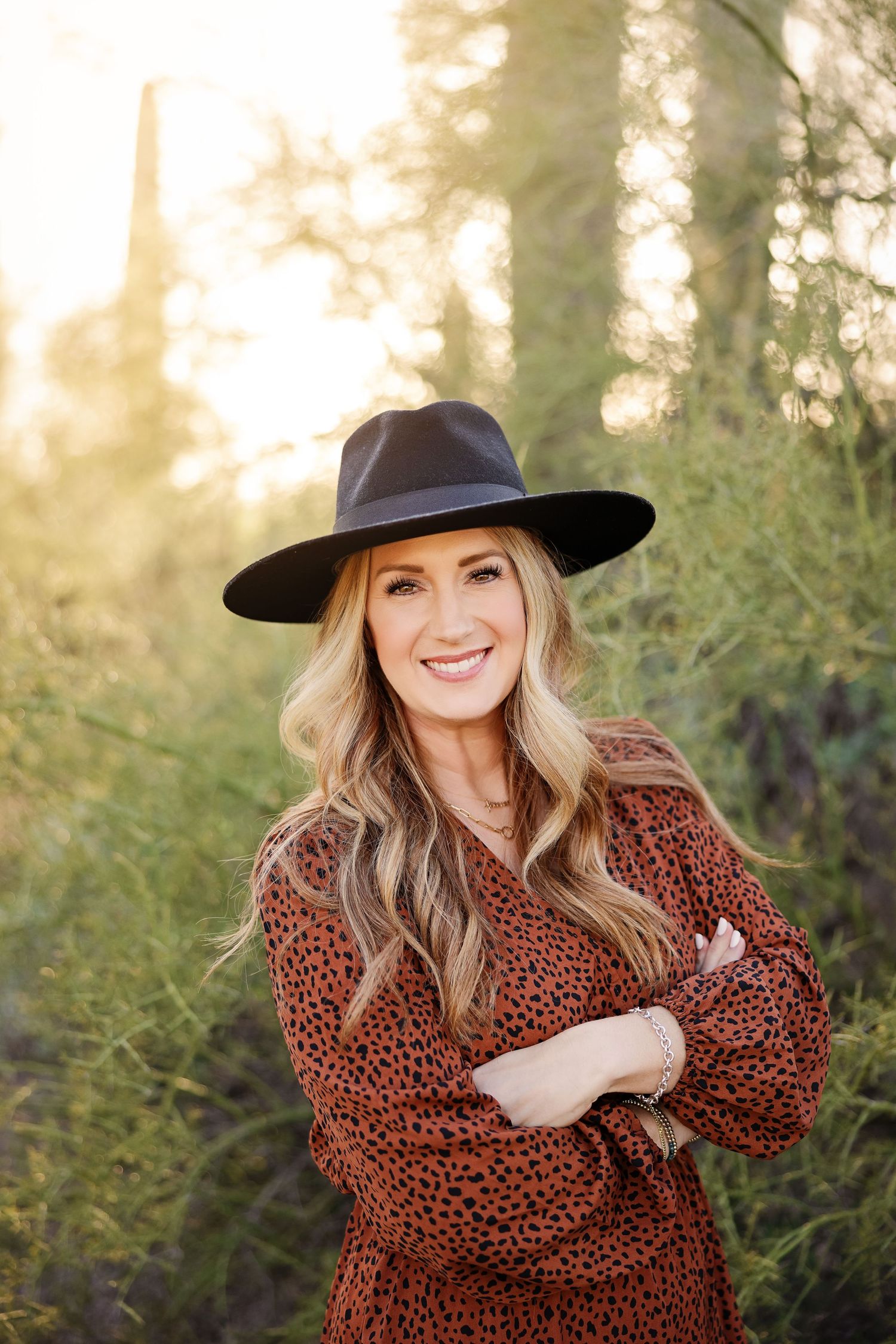 Woman in wide brim hat and brown dress poses confidently against sunlit forest background.