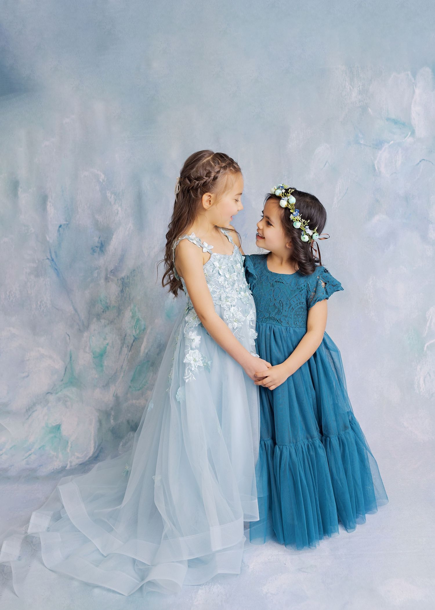 Two girls in blue and light blue formal dresses with floral details standing together against a textured backdrop.