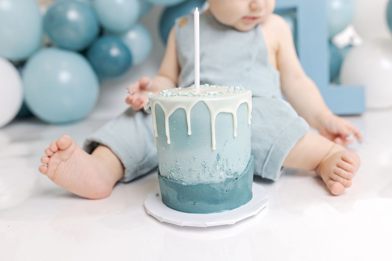 Close up of light blue drip cake with white frosting and single candle for first birthday celebration.