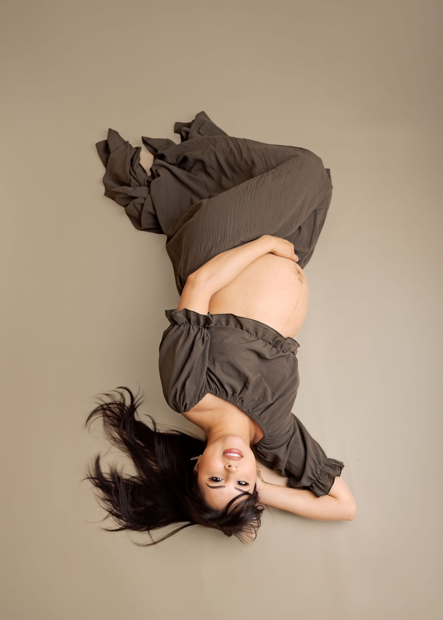 Pregnant woman lying on her back on a beige studio floor, wearing olive green wide-leg pants, cradling her bare baby bump.
