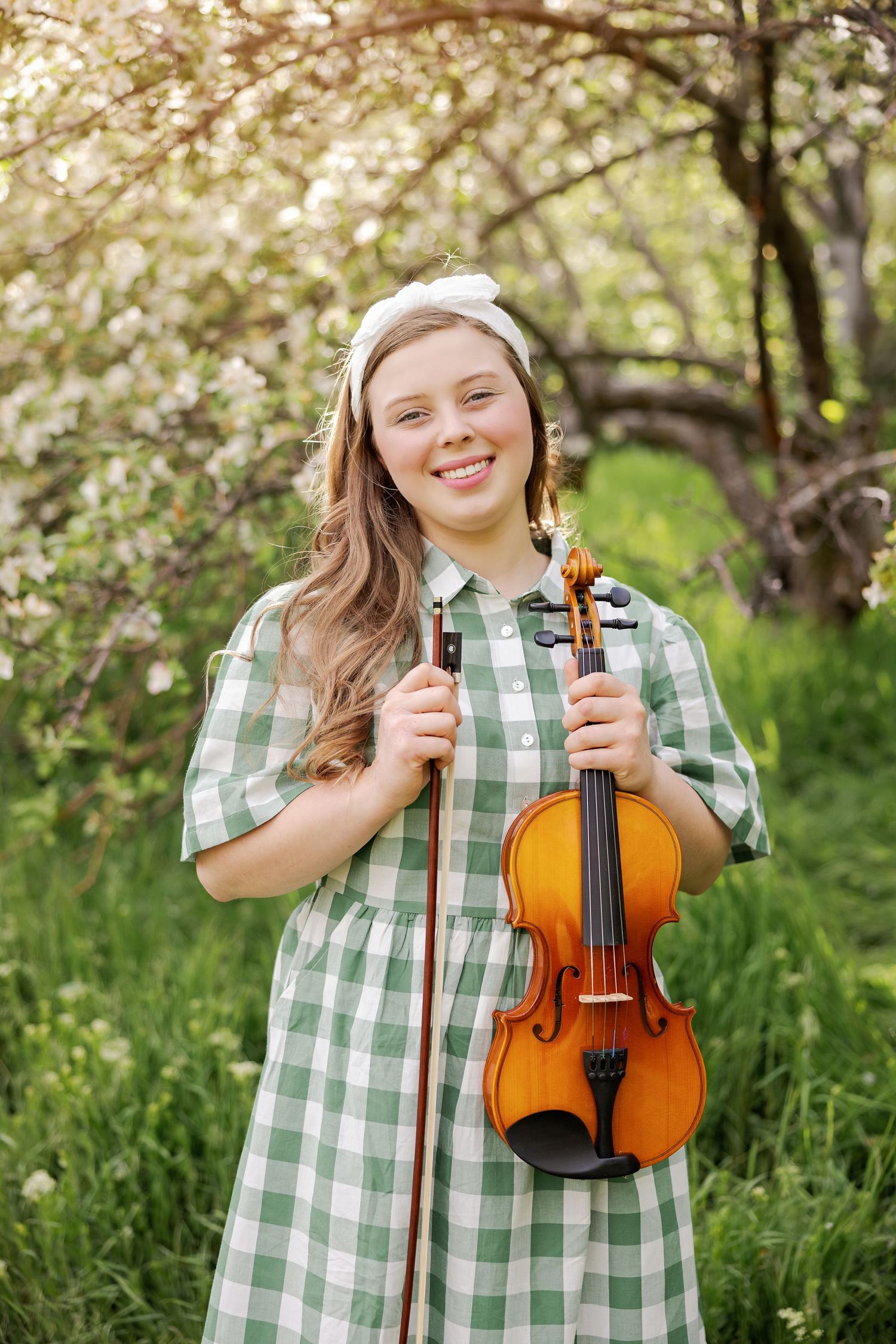 Musician in green gingham dress poses with violin among blooming spring trees.