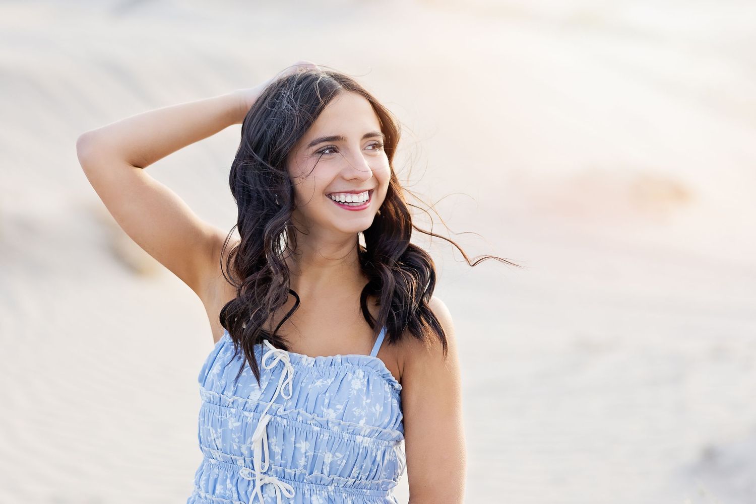 Someone wearing a light blue summer dress poses outdoors with wind blowing through their long dark wavy hair.