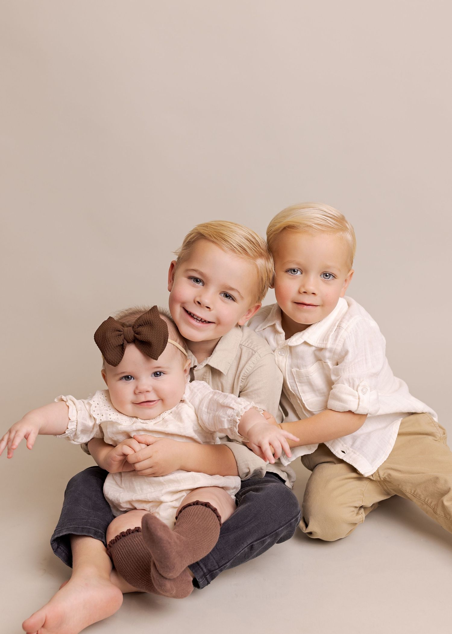 Three young children in coordinating cream and beige outfits sitting together for a studio portrait.