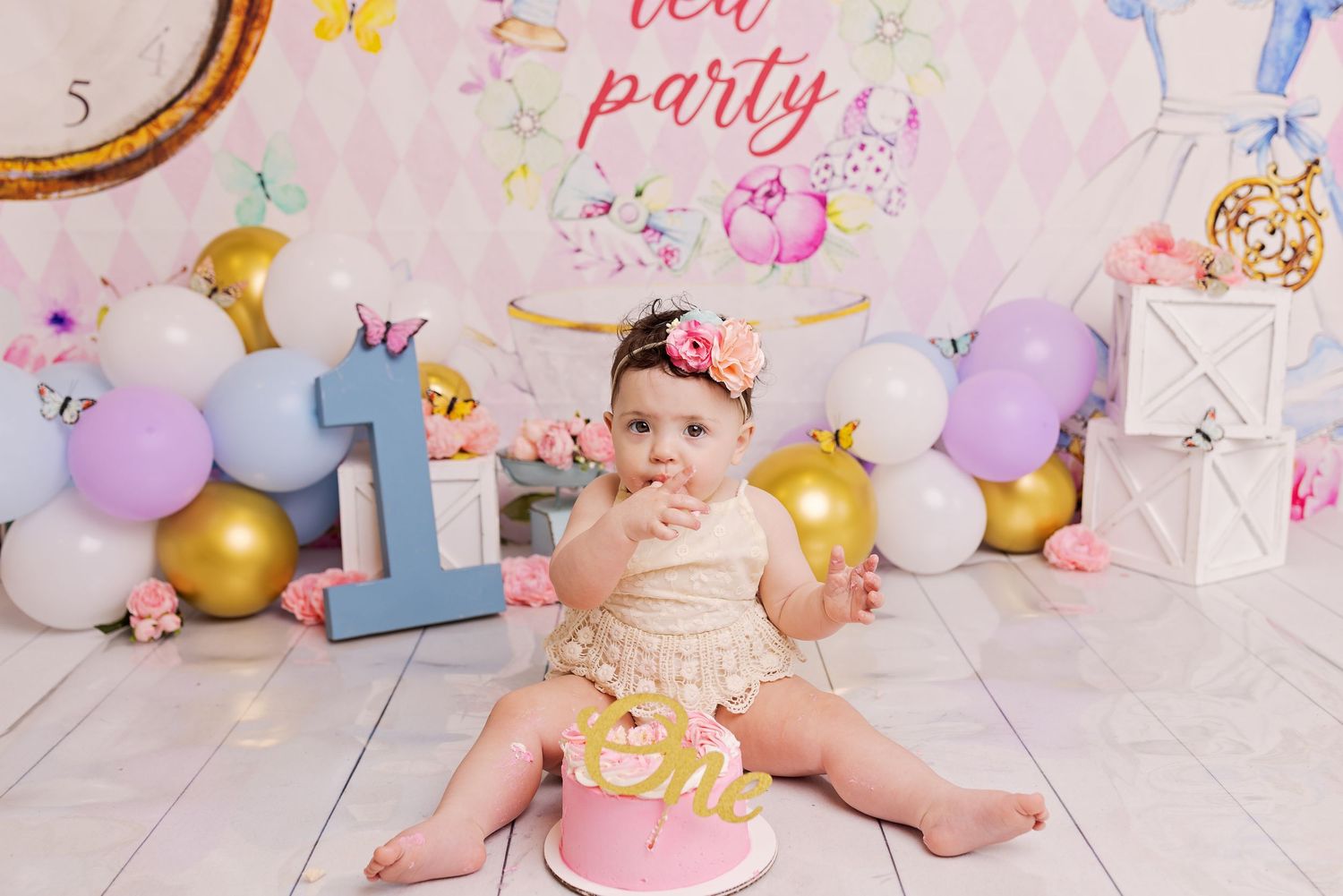 First birthday party setup with pink and purple balloons, sparkly decorations, and a smash cake against a white backdrop.