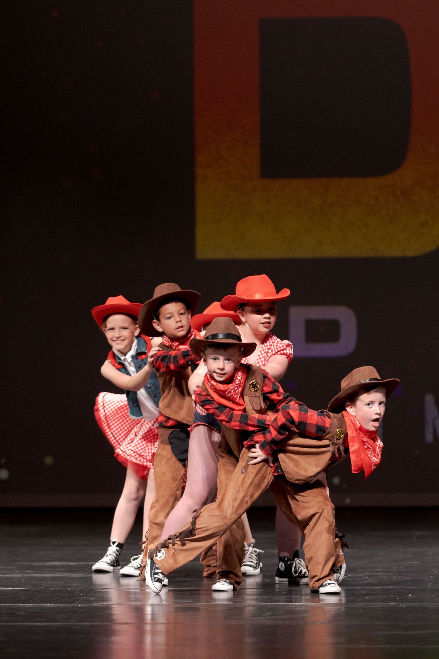 Young dancers perform a western-themed dance routine on stage wearing cowboy hats and matching red costumes.