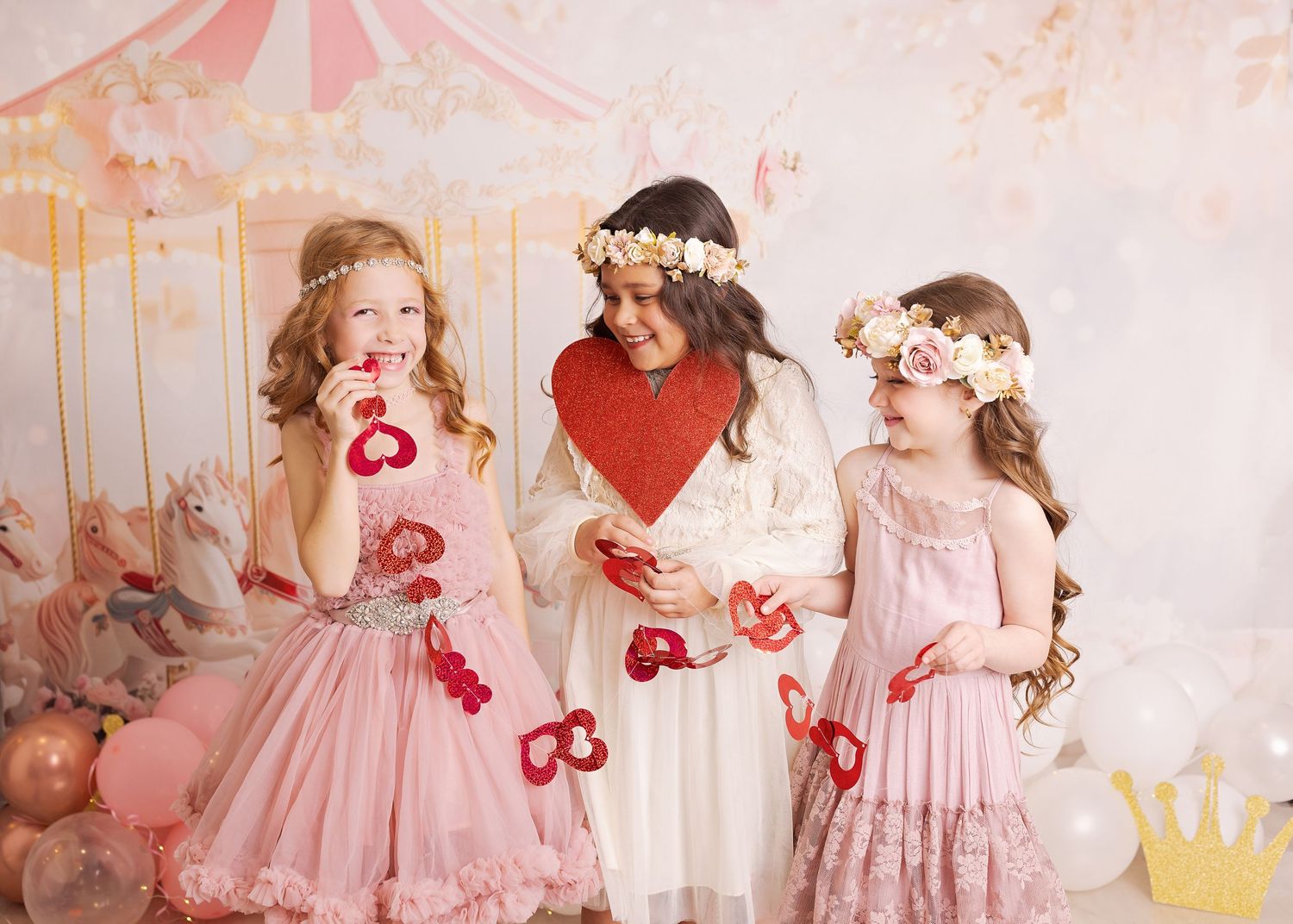 Three people in vintage-style dresses with floral crowns pose together against a pink and peach backdrop with paper decorations.