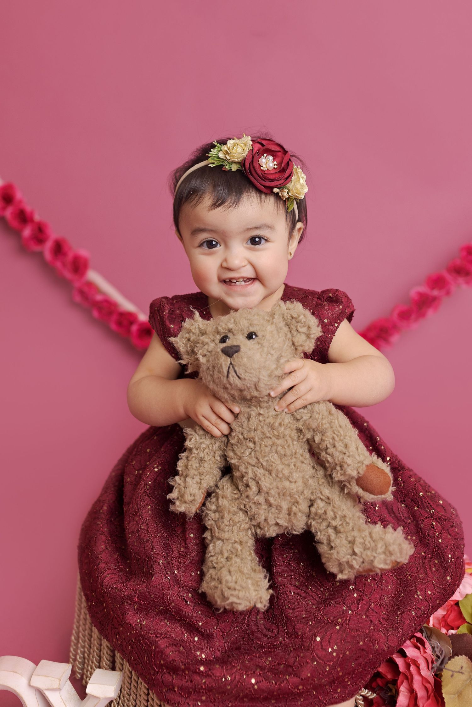 A child in a sparkly burgundy dress poses with a plush teddy bear against a pink background.