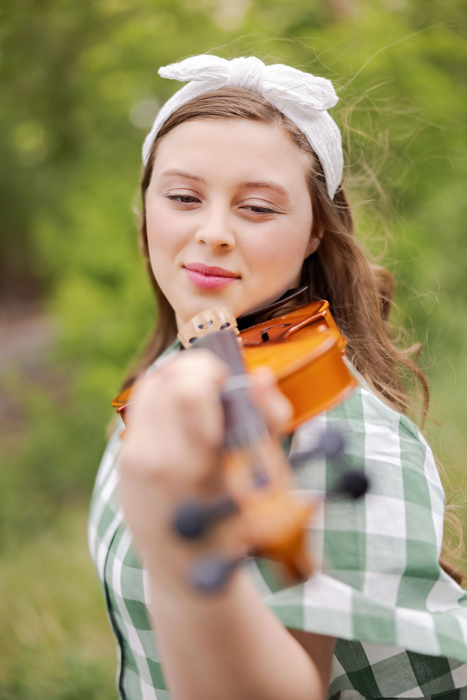 Musician in green gingham dress and white headband plays violin outdoors.