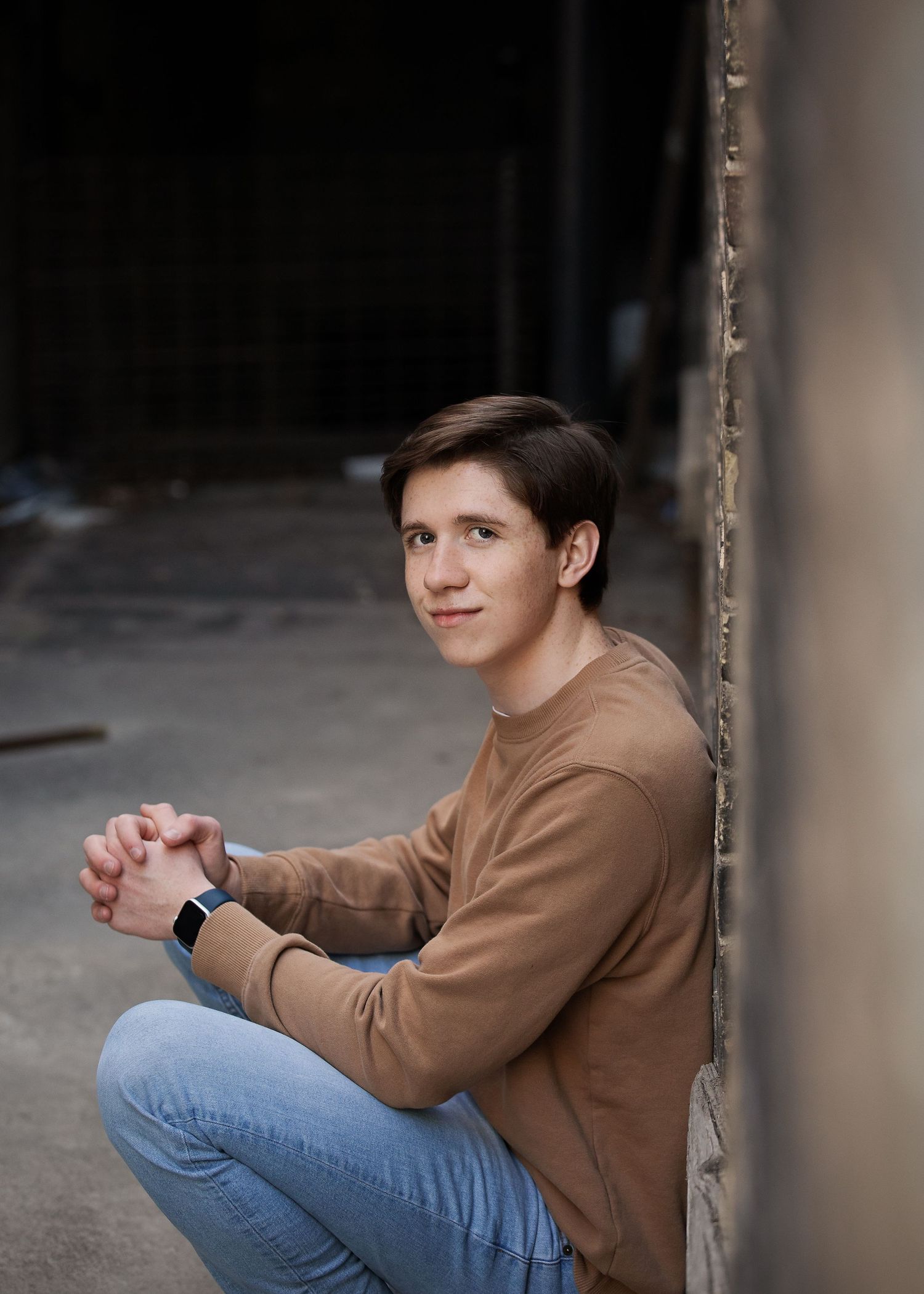 Casual portrait in tan sweater and jeans sitting against concrete wall.