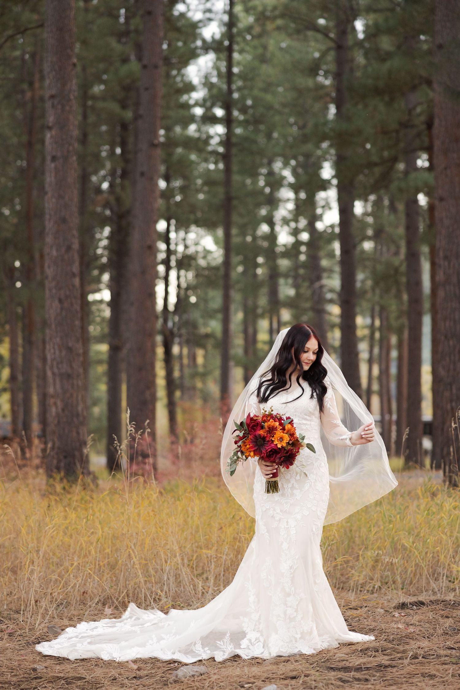 Elegant lace wedding dress with long train and fall-colored bouquet photographed in a pine forest setting.