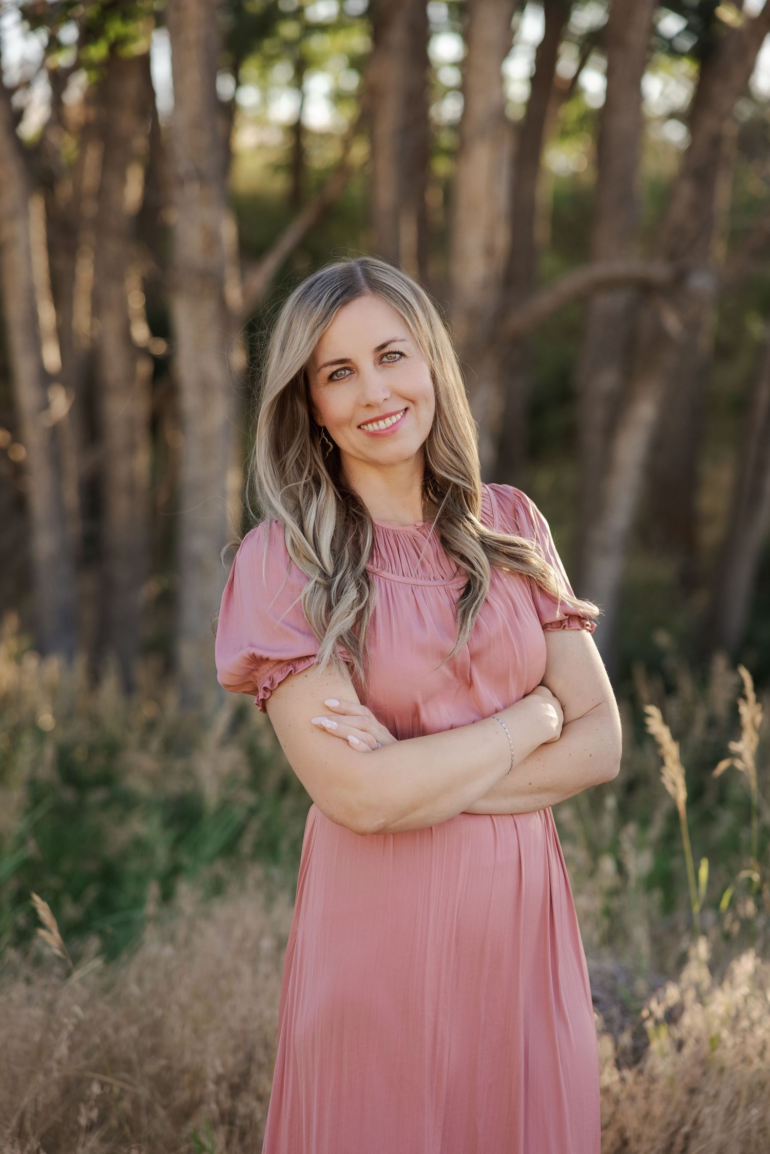 Woman in pink dress stands confidently in front of wooded area during golden hour.