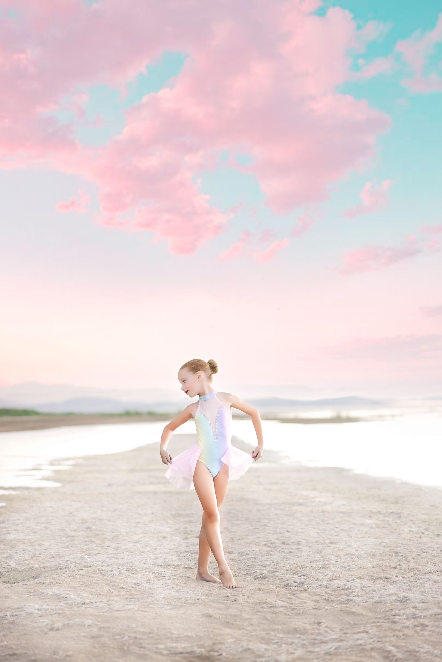 A young dancer in white leotard poses gracefully on a sandy beach under pink and turquoise sunset skies.